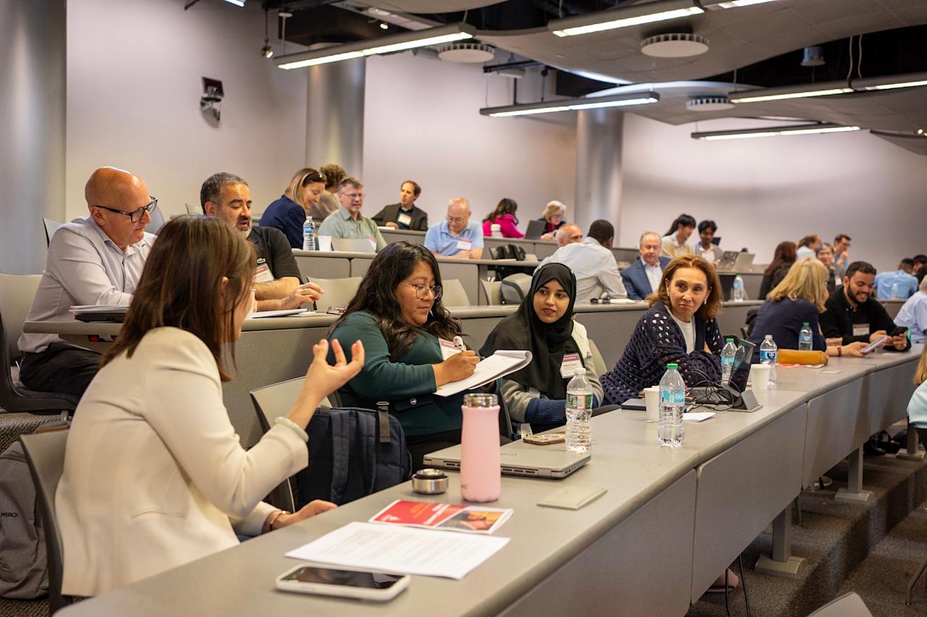 A row of four women sitting in the same row in a lecture hall having a conversation and making hand gestures while being engaged in conversation with each other.