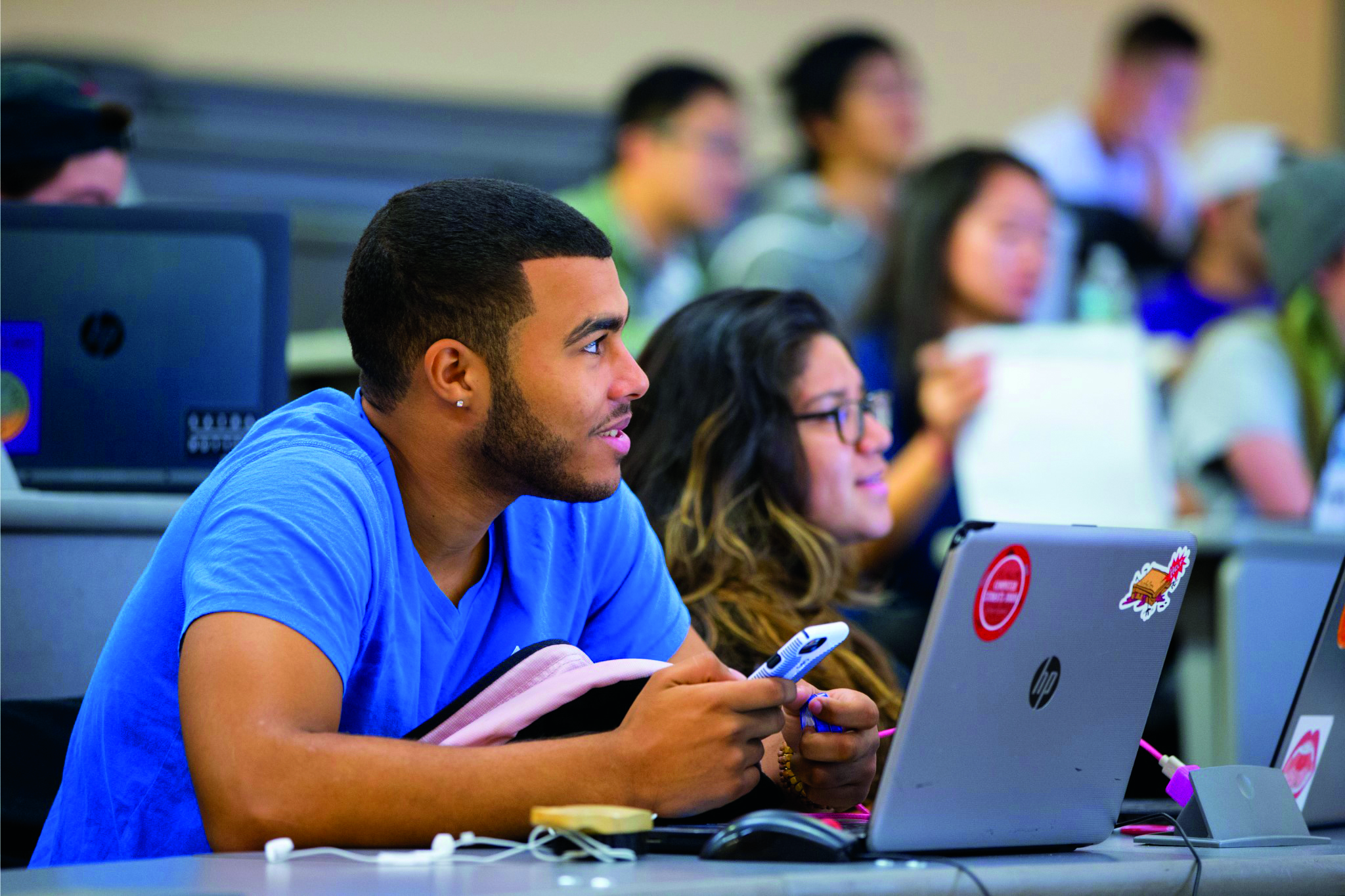 A black, college-aged man sits at a desk, listening to a lecture. He’s holding a cell phone in his hands and a laptop sits open in front of him.