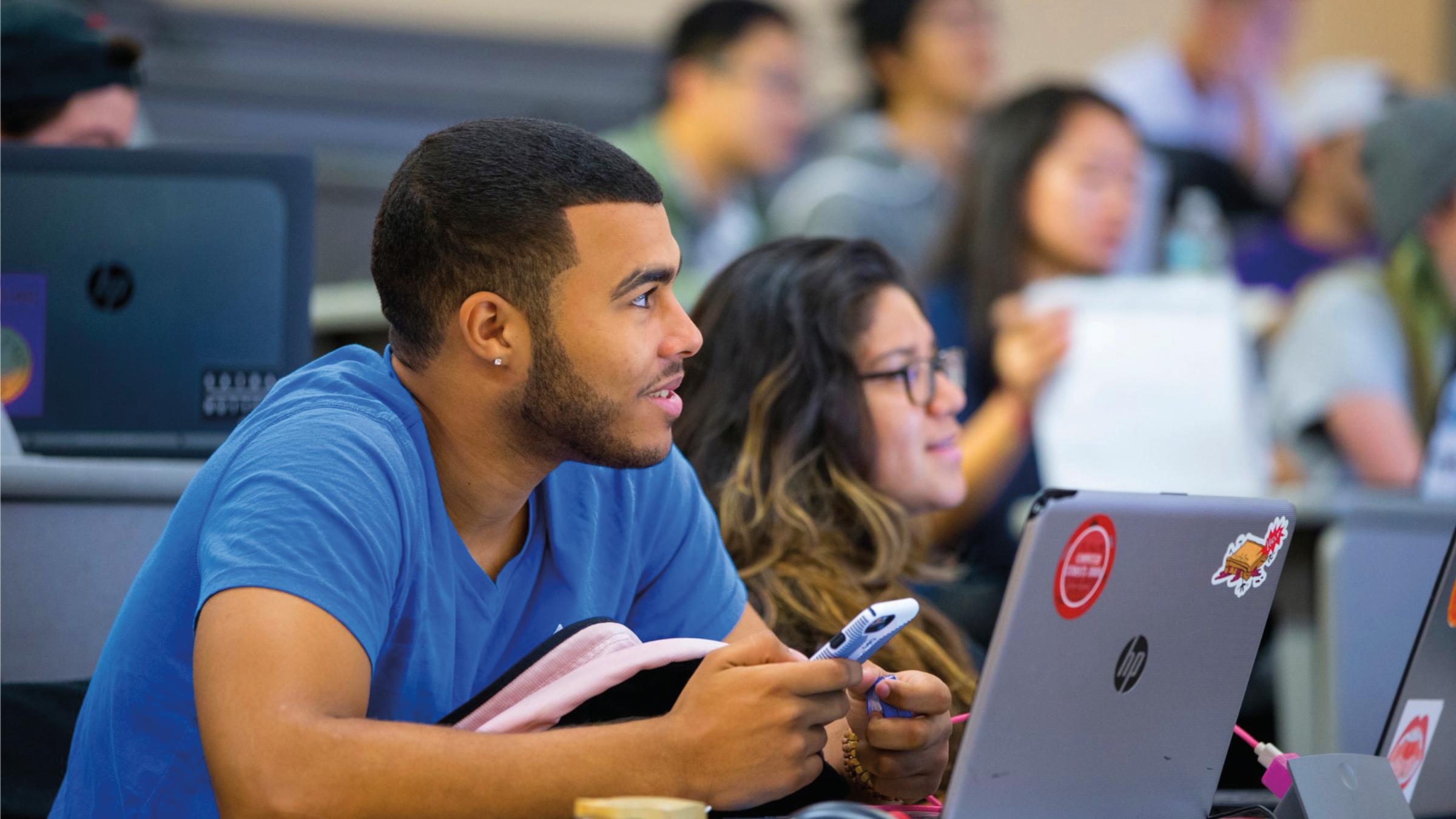 A black, college-aged man sits at a desk, listening to a lecture. He’s holding a cell phone in his hands and a laptop sits open in front of him.