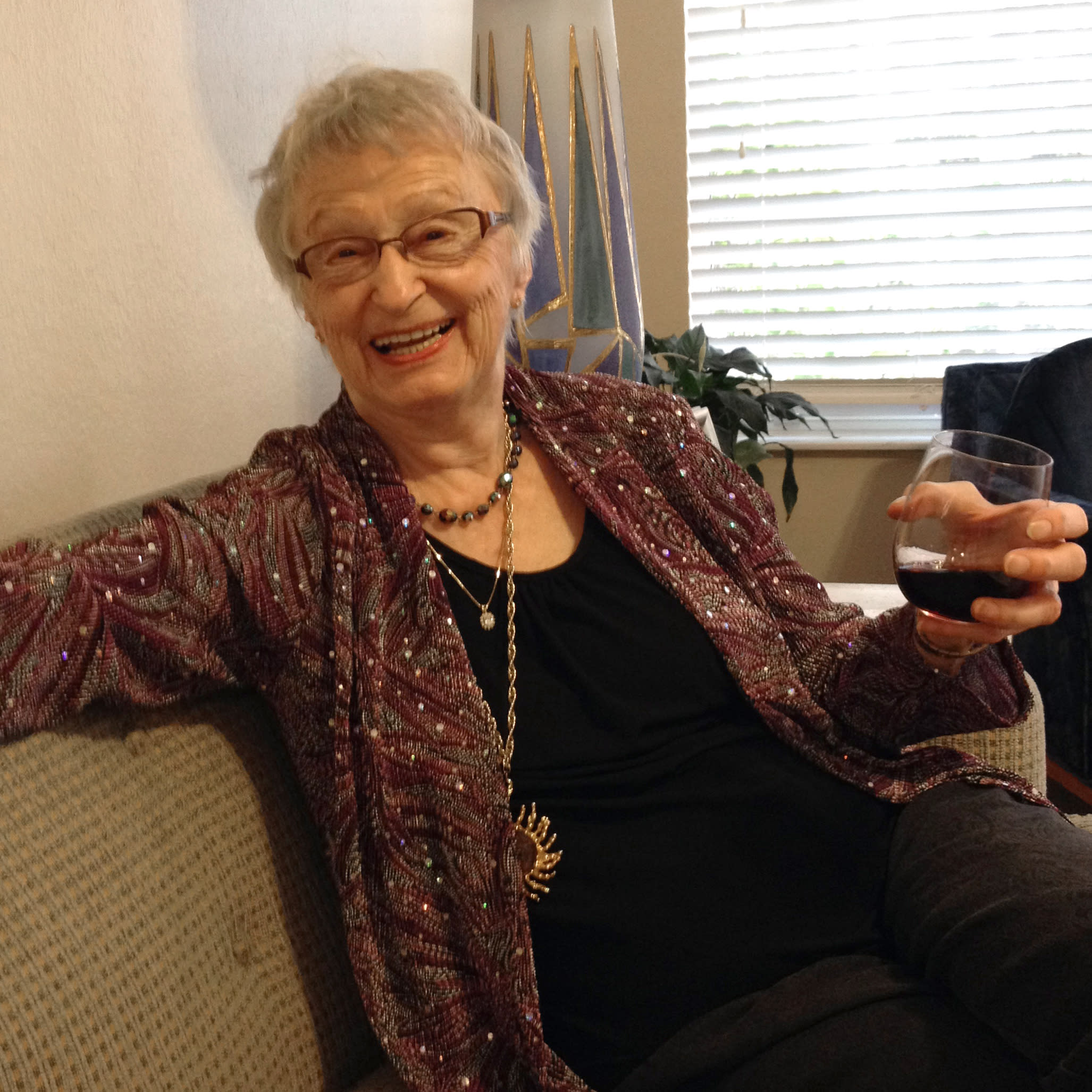 Photo of an older woman seated on a couch, smiling and holding a glass of wine.