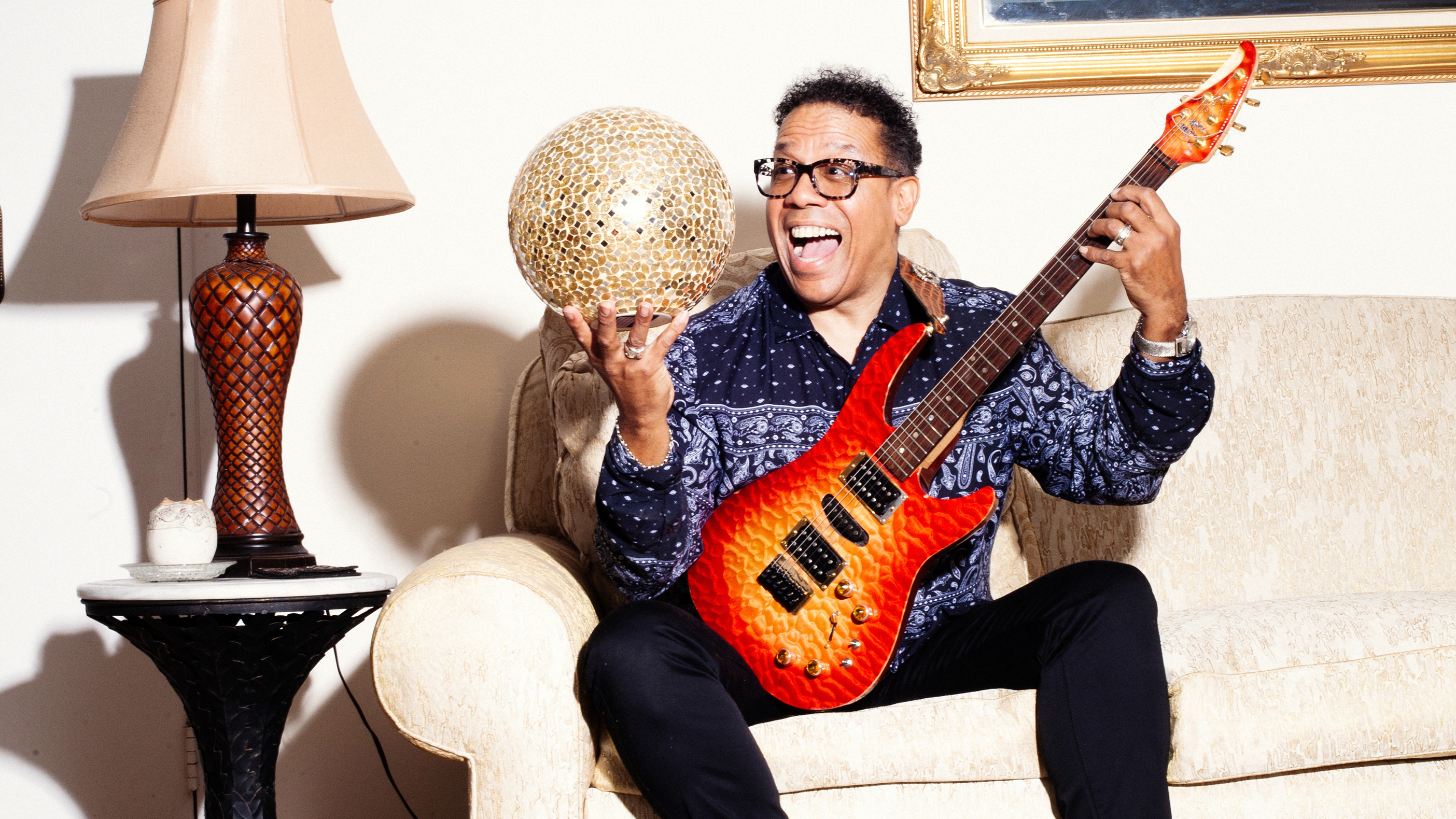 Carlos Alomar joyfully sits on a couch with his guitar and a golden orb.