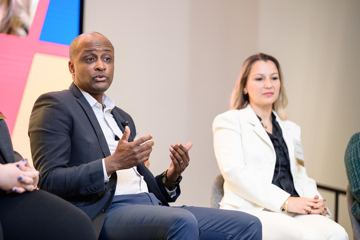 Panelists participate in a discussion onstage at a professional event. One speaker in a dark suit gestures while speaking, while another panelist in a light-colored suit listens.