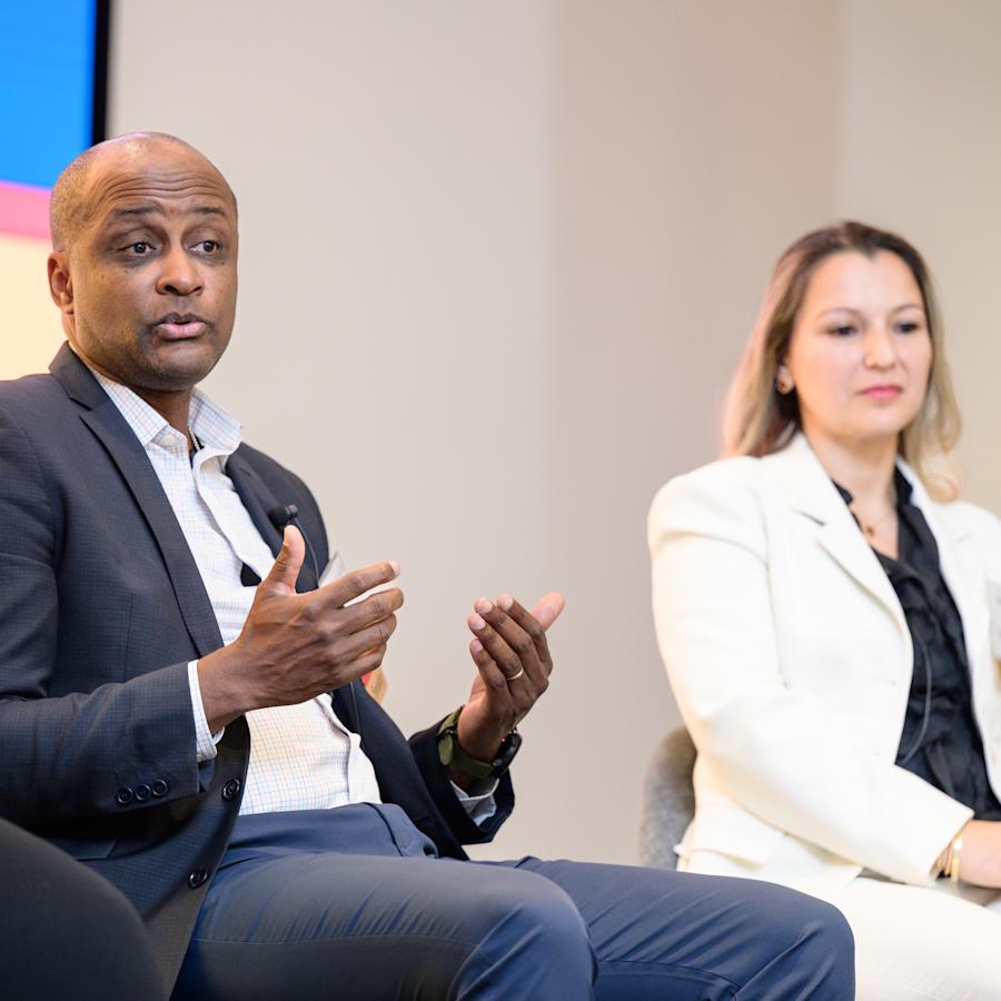 Panelists participate in a discussion onstage at a professional event. One speaker in a dark suit gestures while speaking, while another panelist in a light-colored suit listens.