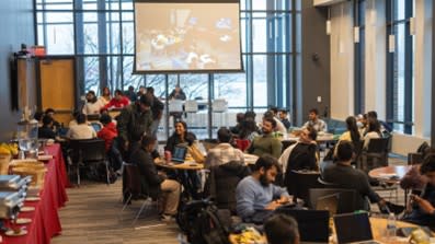 A room full of students sitting with their laptops and lunches. There is a long table of catered food on the left.