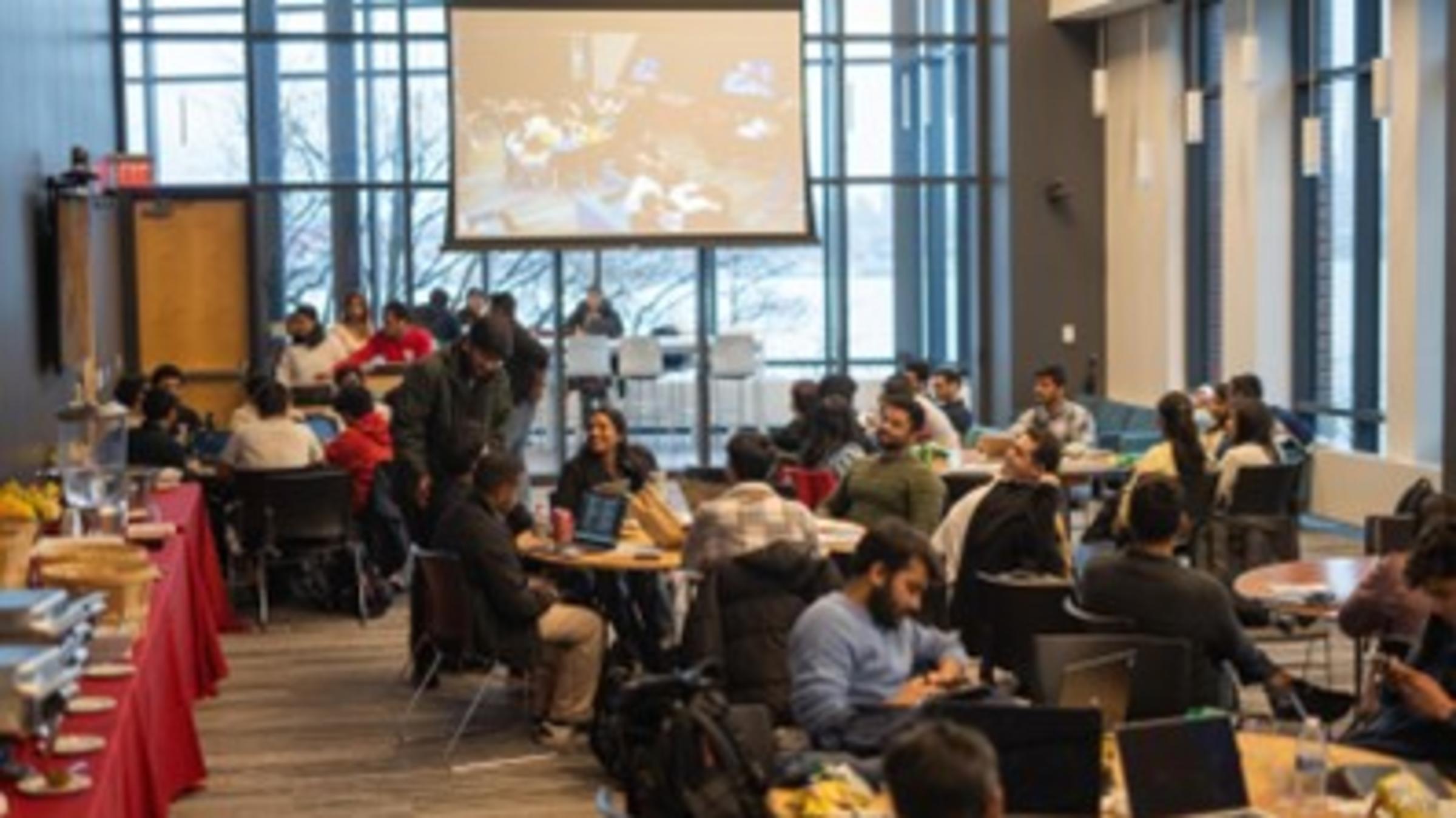 A room full of students sitting with their laptops and lunches. There is a long table of catered food on the left.