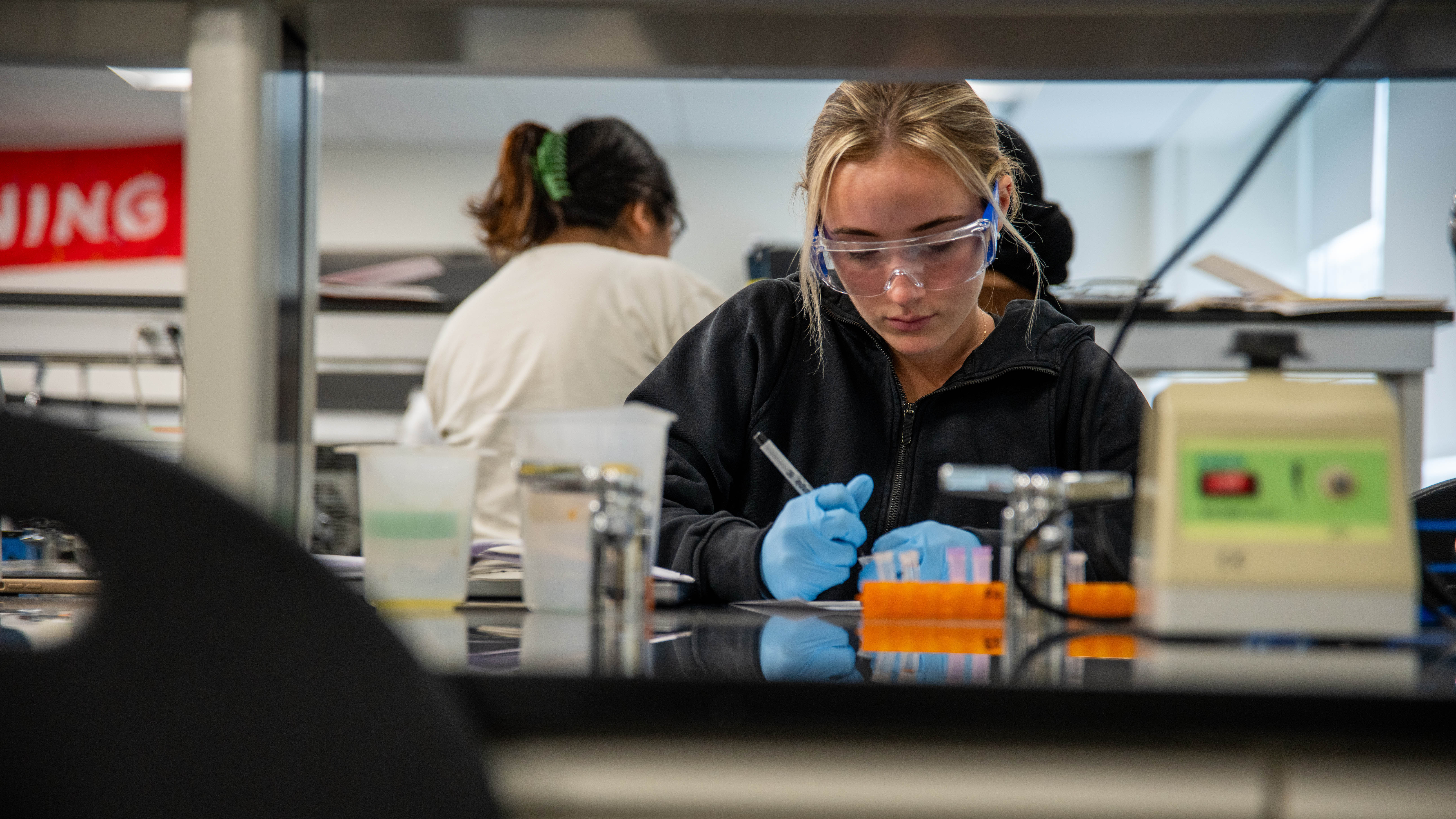 Student in a lab wearing safety goggles and writing notes.