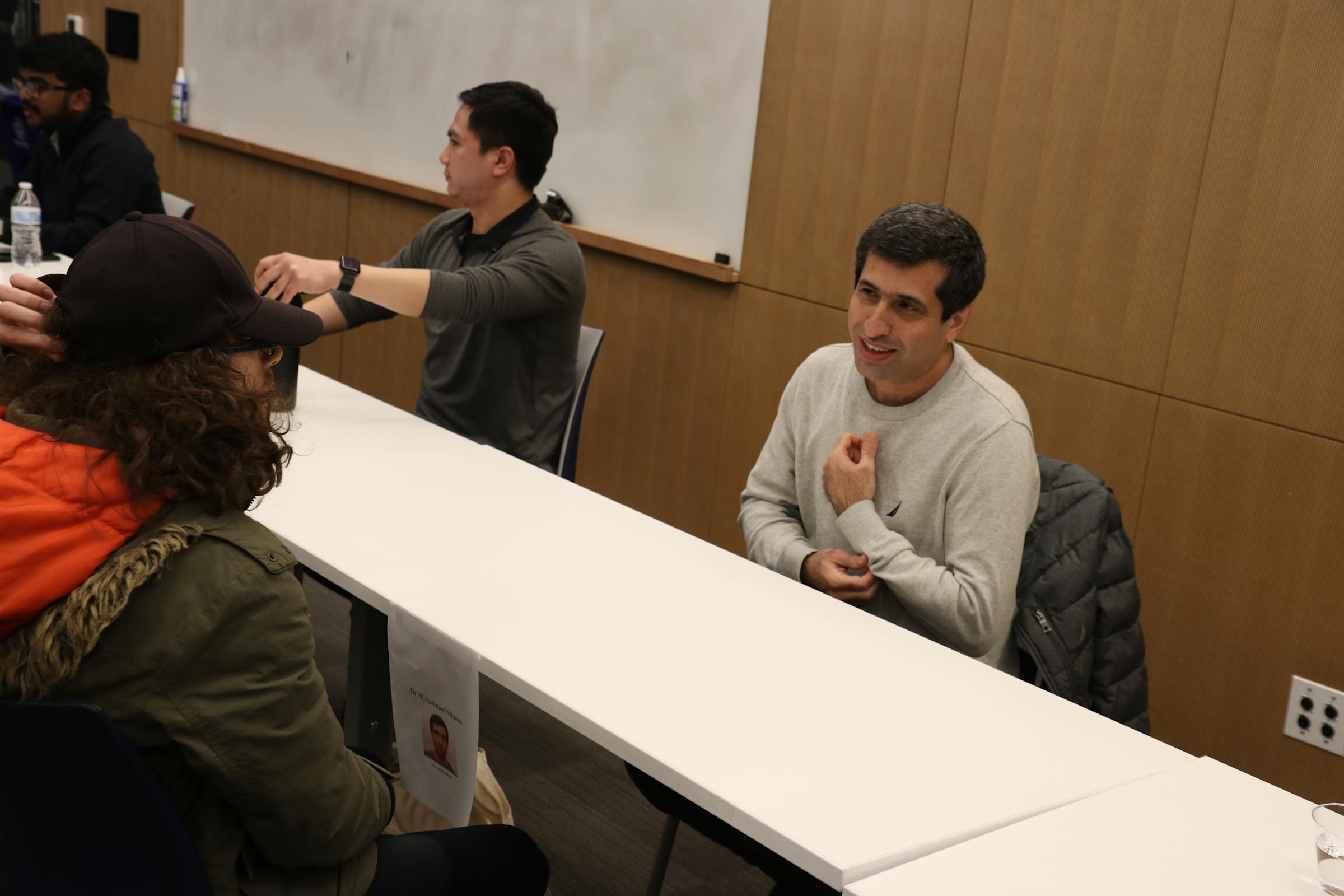 People sit across a table on both sides facing each other in conversation. Some are students, some are industry professionals.