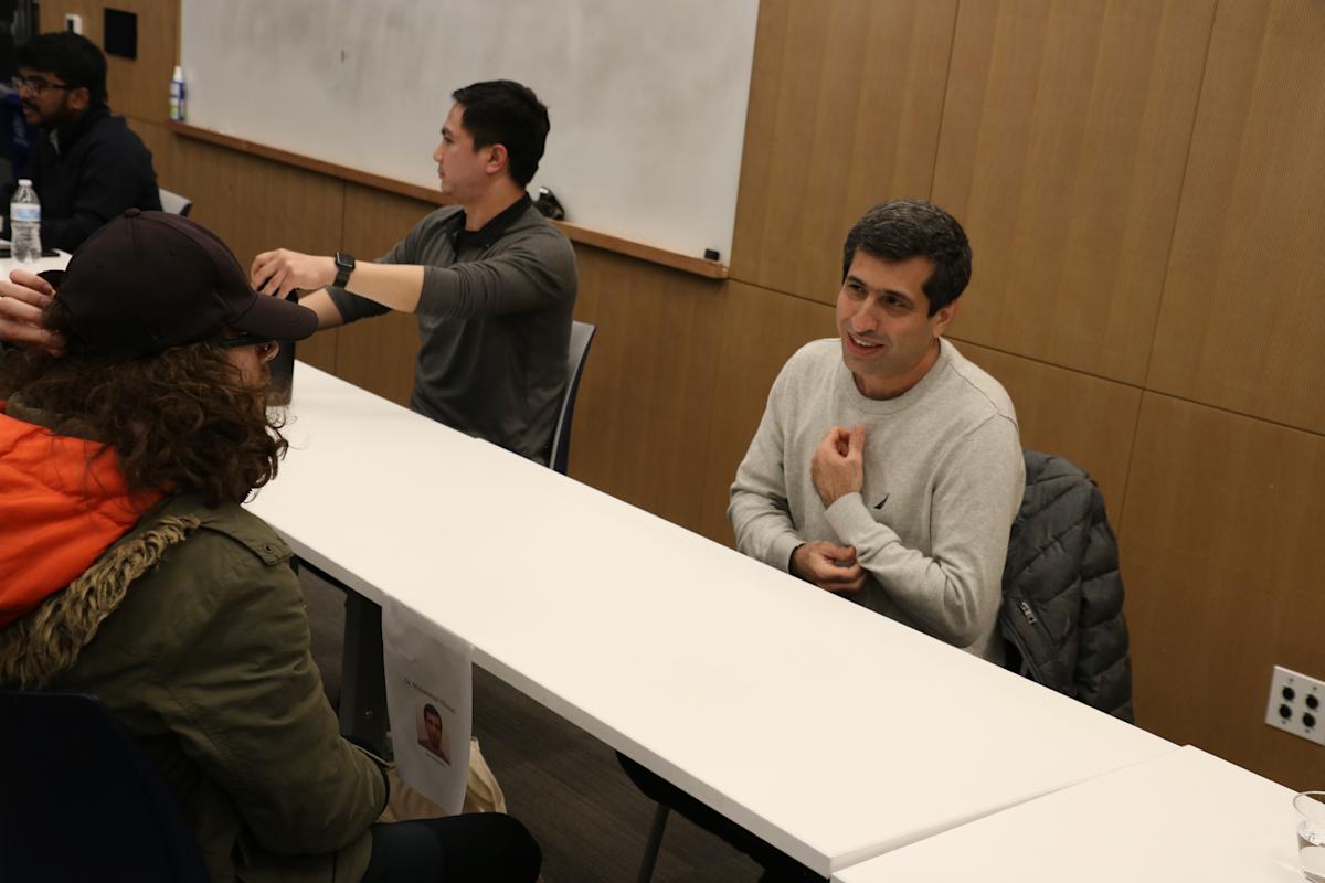 People sit across a table on both sides facing each other in conversation. Some are students, some are industry professionals.