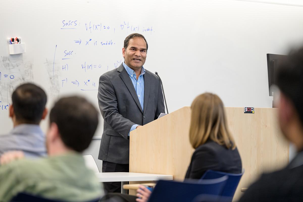 A professor speaking at a podium, gesturing to the audience.