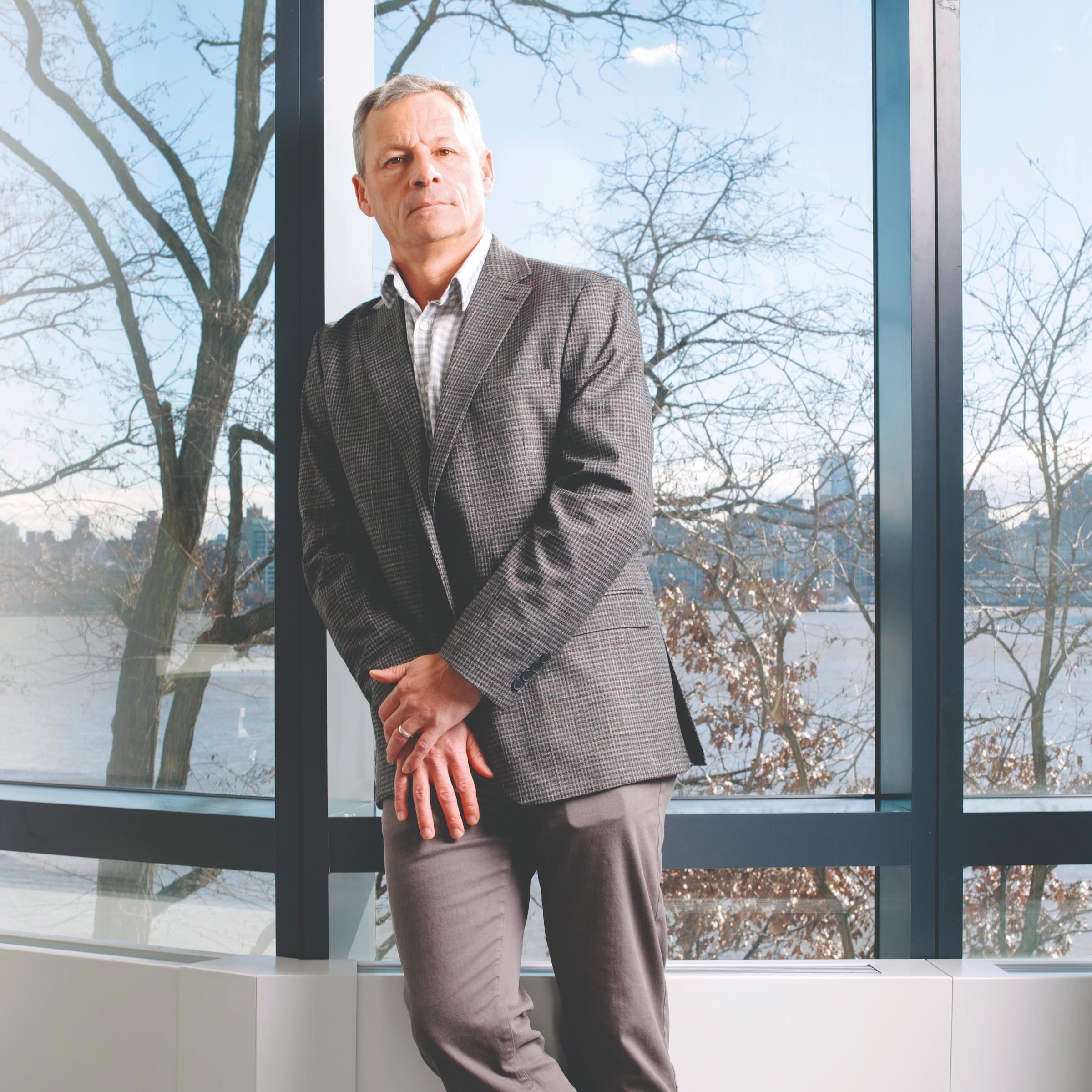 Matthew Ketschke, Stevens Class of 1995 and 1998, leans his right shoulder against the sill of a large window. Behind him, through the window, are bare trees and the gray expanse of the Hudson River.