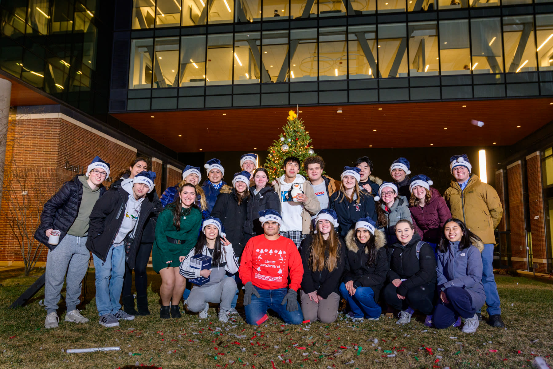 A group of diverse students smiling and posing together in front of a beautifully decorated Christmas tree on campus. The students are wearing festive hats, and the tree is adorned with lights, ornaments and a rubber duck on top.
