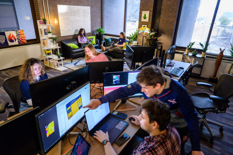Students collaborate at computer workstations in a bright campus innovation space, while others work on laptops in a lounge area with couches, whiteboards and large windows.