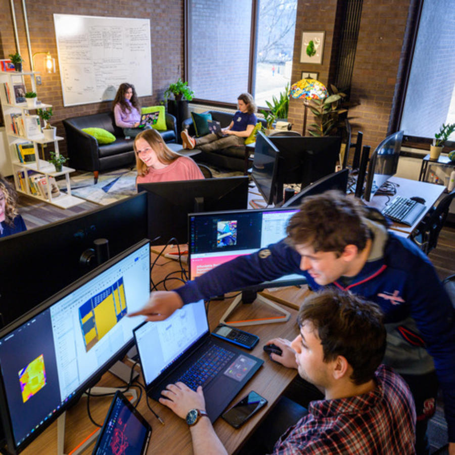 Students collaborate at computer workstations in a bright campus innovation space, while others work on laptops in a lounge area with couches, whiteboards and large windows.