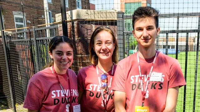 Three students smile at a table on campus with Stevens Ducks shirts on