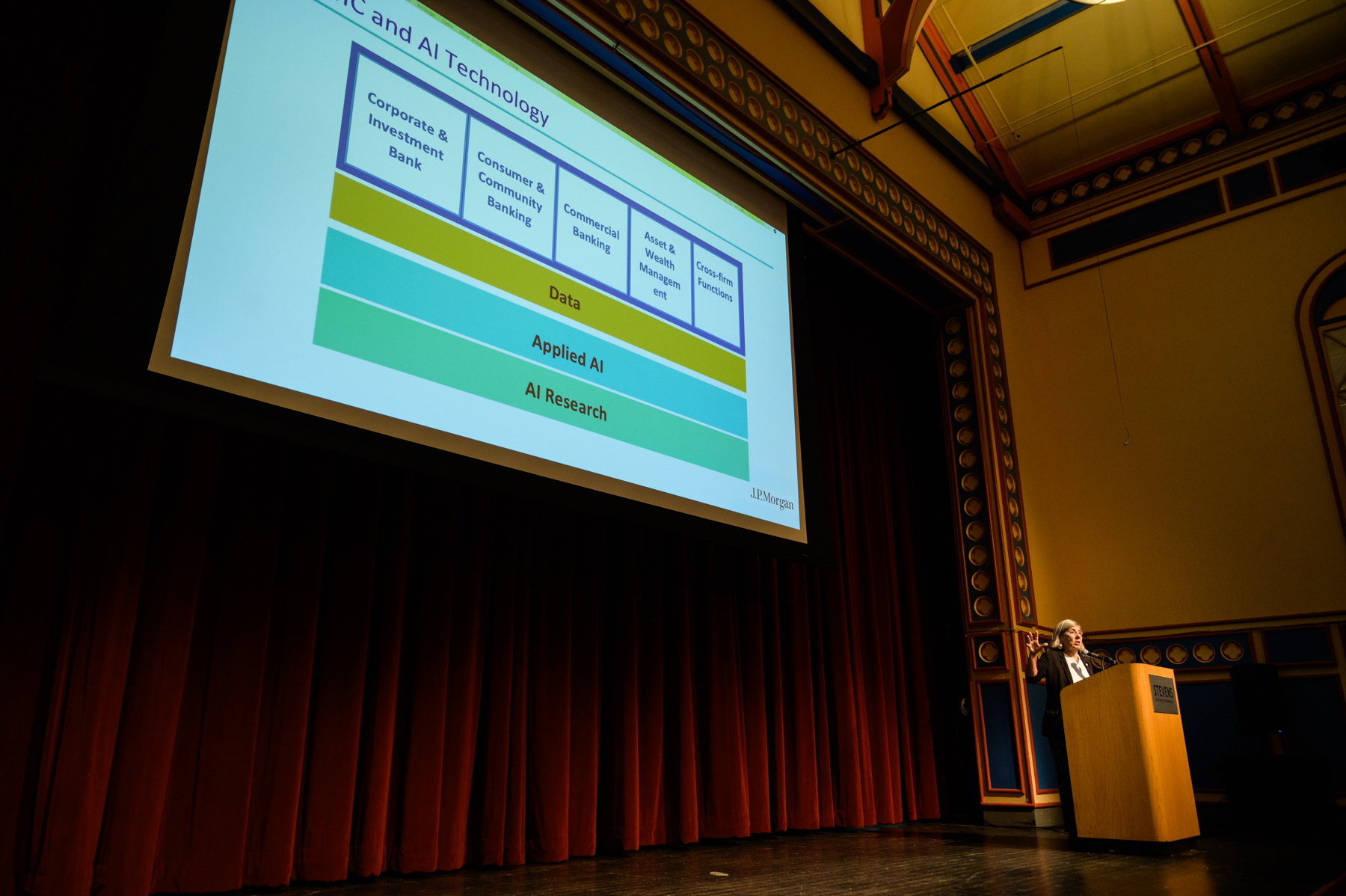 Manuela Veloso stands at a podium in an auditorium giving a presentation with a large slide displaying a diagram of business divisions supported by layers labeled “Data,” “Applied AI,” and “AI Research” projected above.  