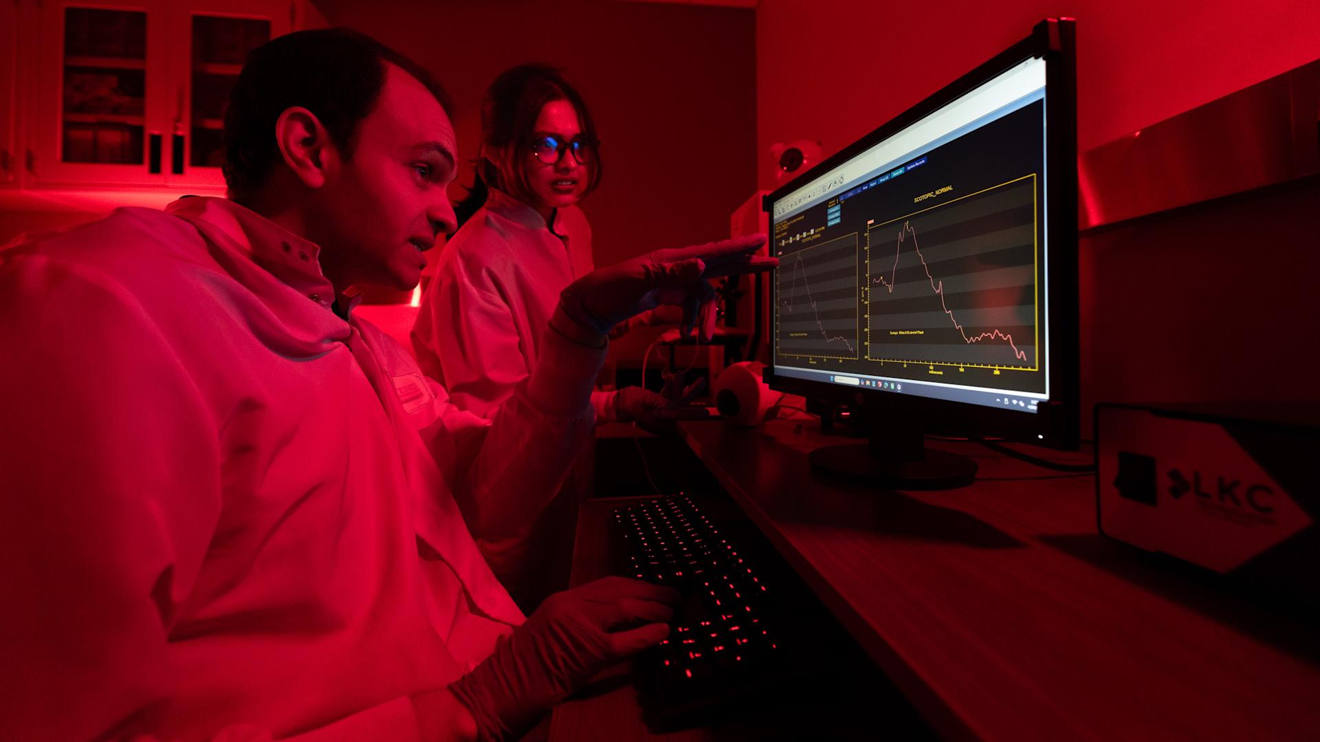 Two students in lab coats looking at a monitor in a dark room lit in red.