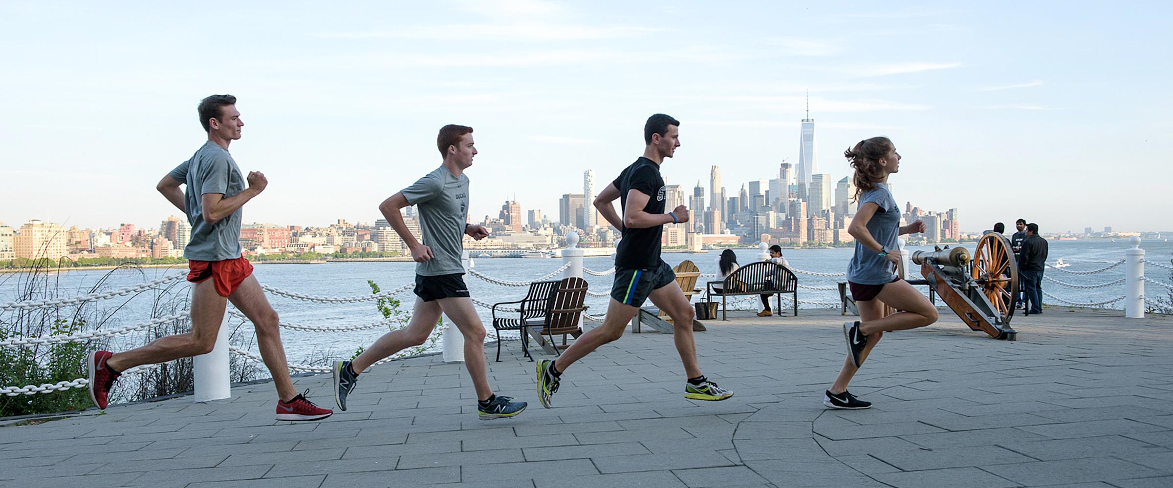 Group of four runners jog past Castle Point Terrace with New York skyline in background.