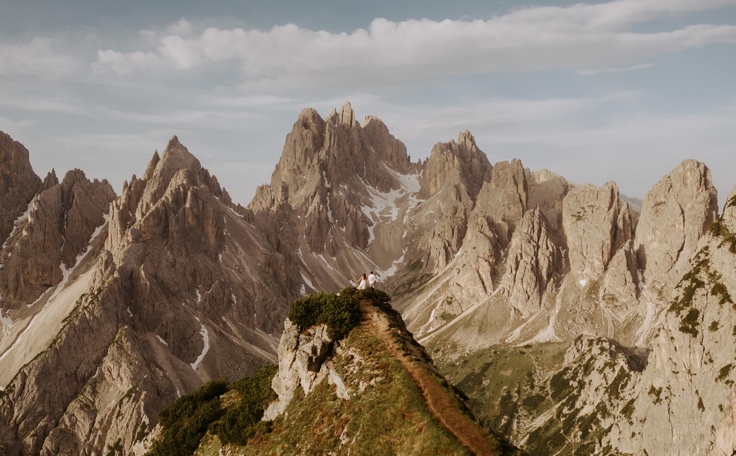 A man and a woman are seen in the distance standing high atop a rocky mountain, surrounded by the high rocky peaks of the mountain range.
