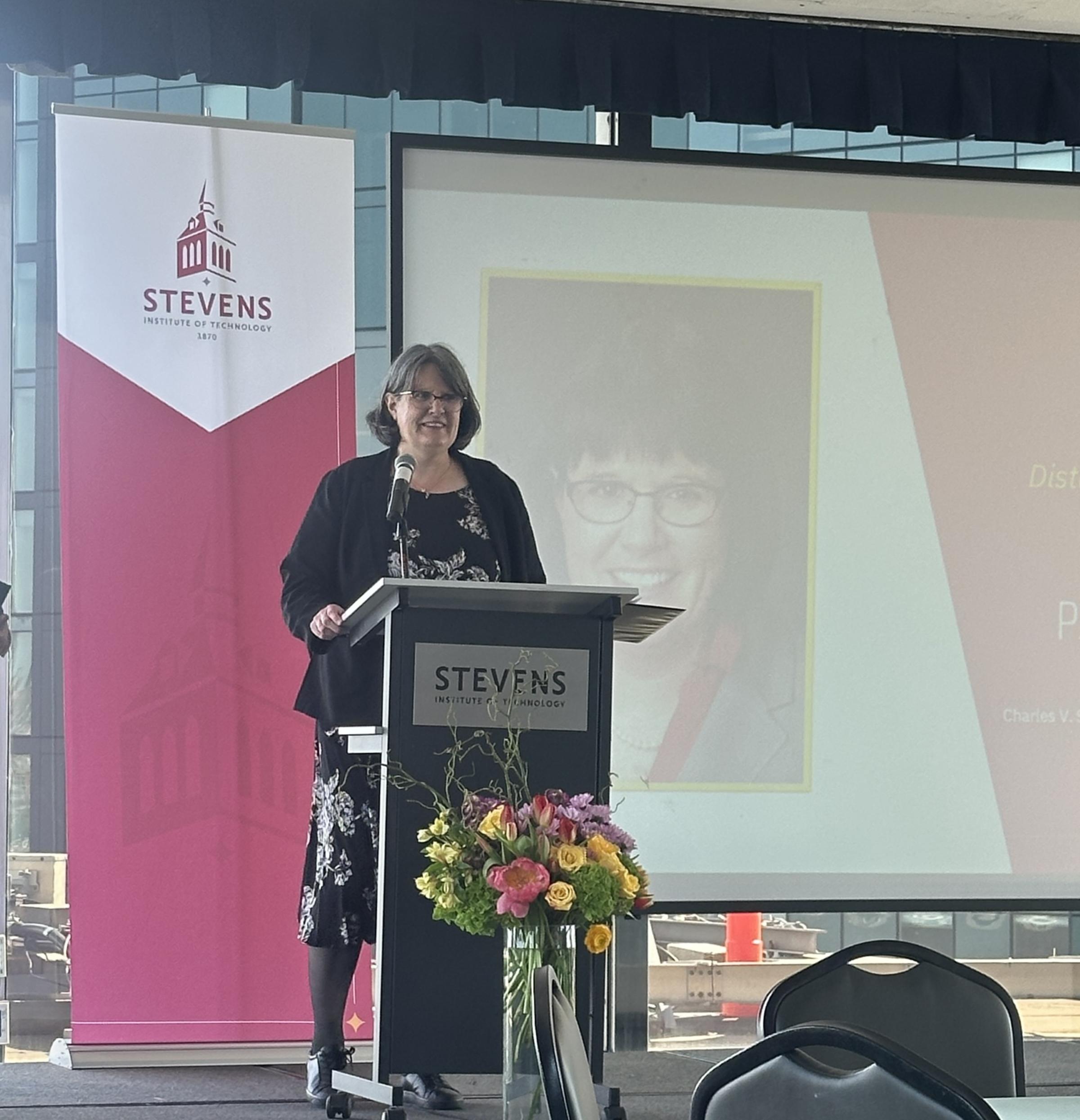 Patricia Muisener stands behind a podium on a stage in front of a presentation and Stevens banner.
