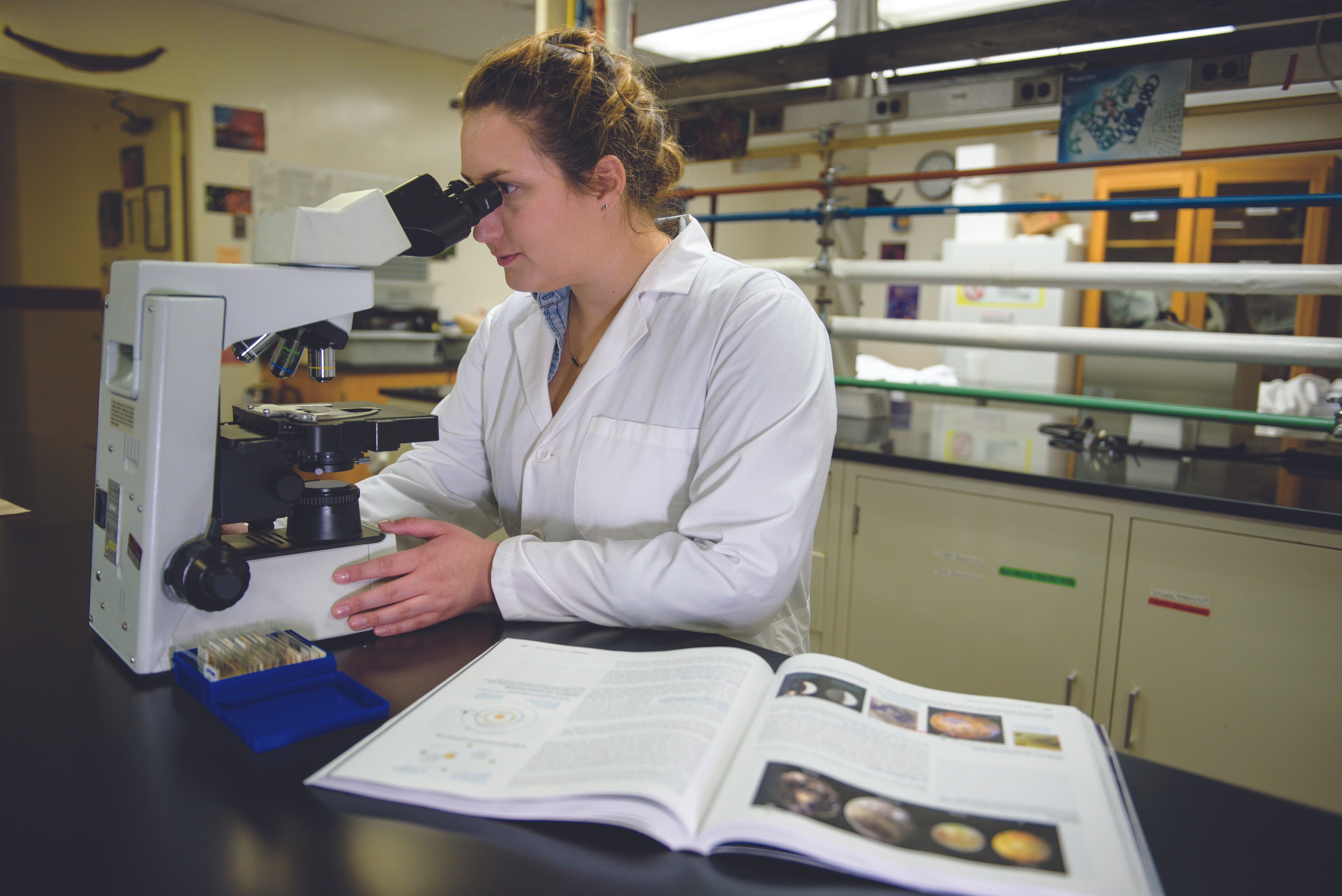 Female student in a lab coat looking in microscope with an open textbook on her right