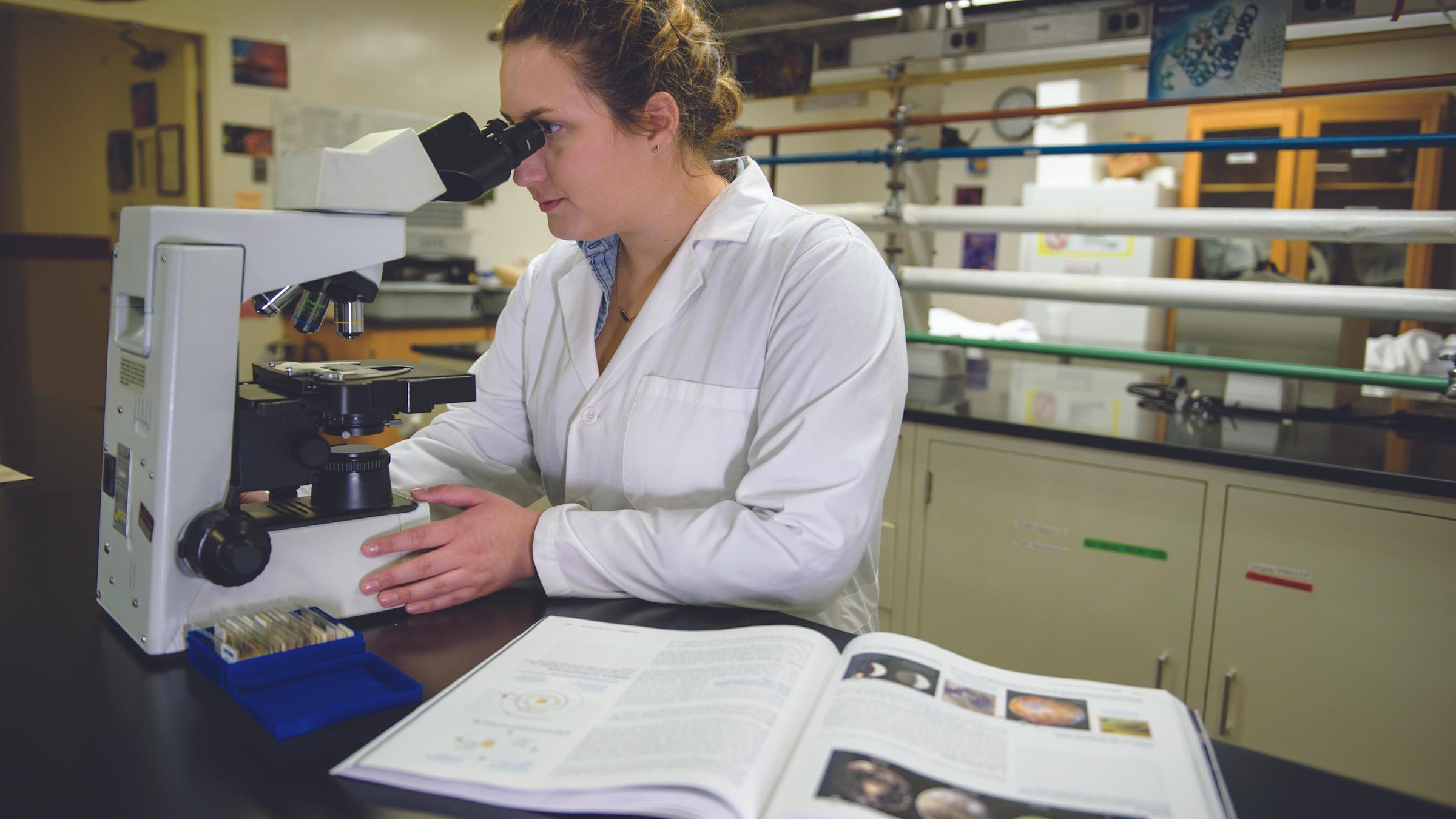 Female student in a lab coat looking in microscope with an open textbook on her right