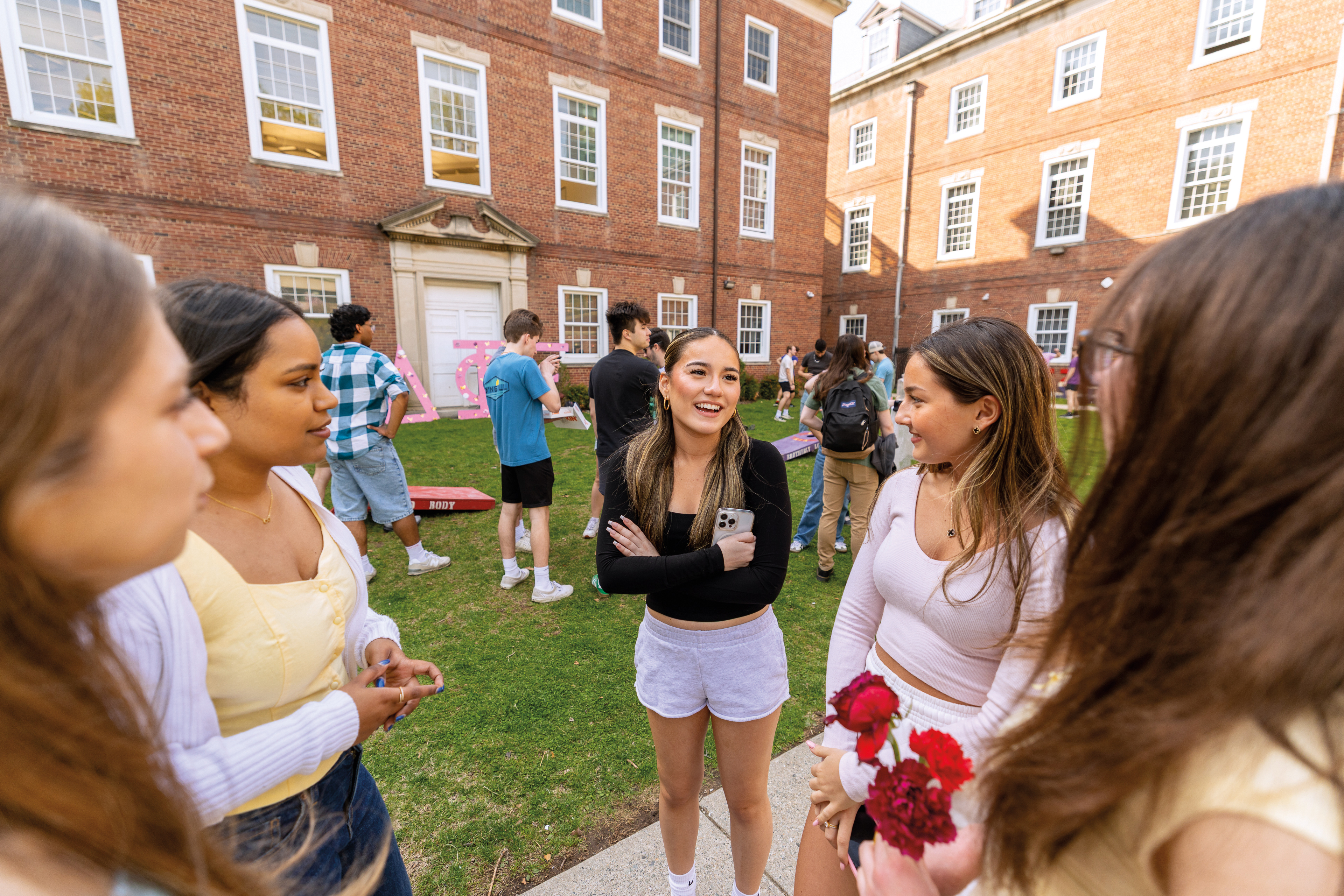 five women students talking in the foreground, with students playing lawn games in the background.