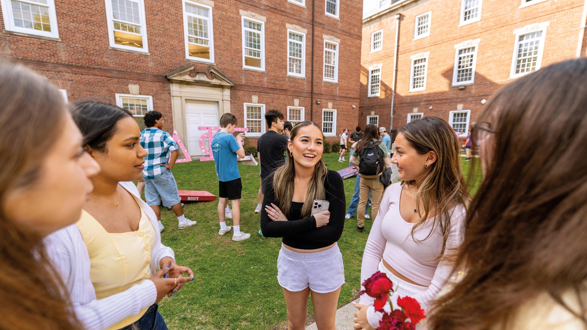 five women students talking in the foreground, with students playing lawn games in the background.