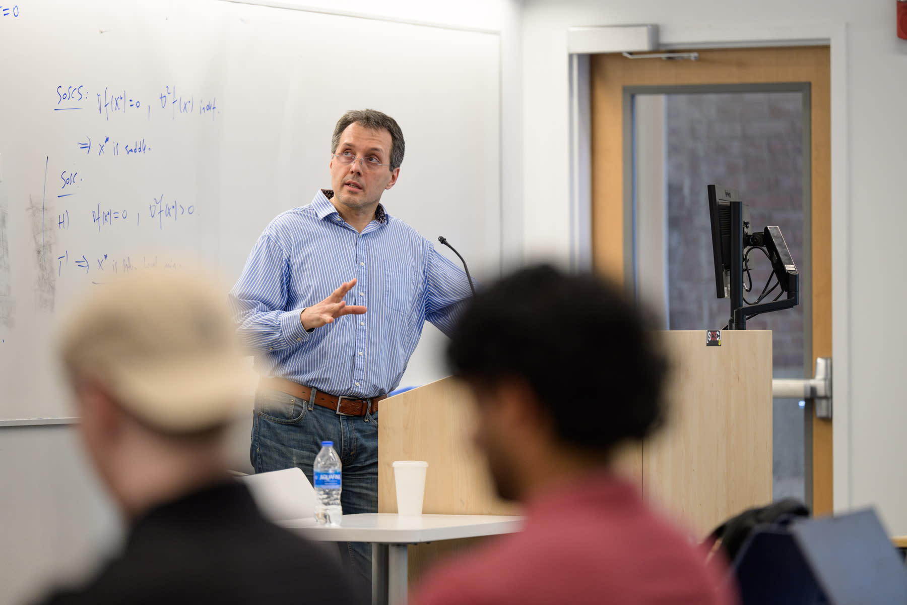 A man standing at a speaking podium, discussing something that is on a screen out of view, to a group of Mathematical Sciences students.
