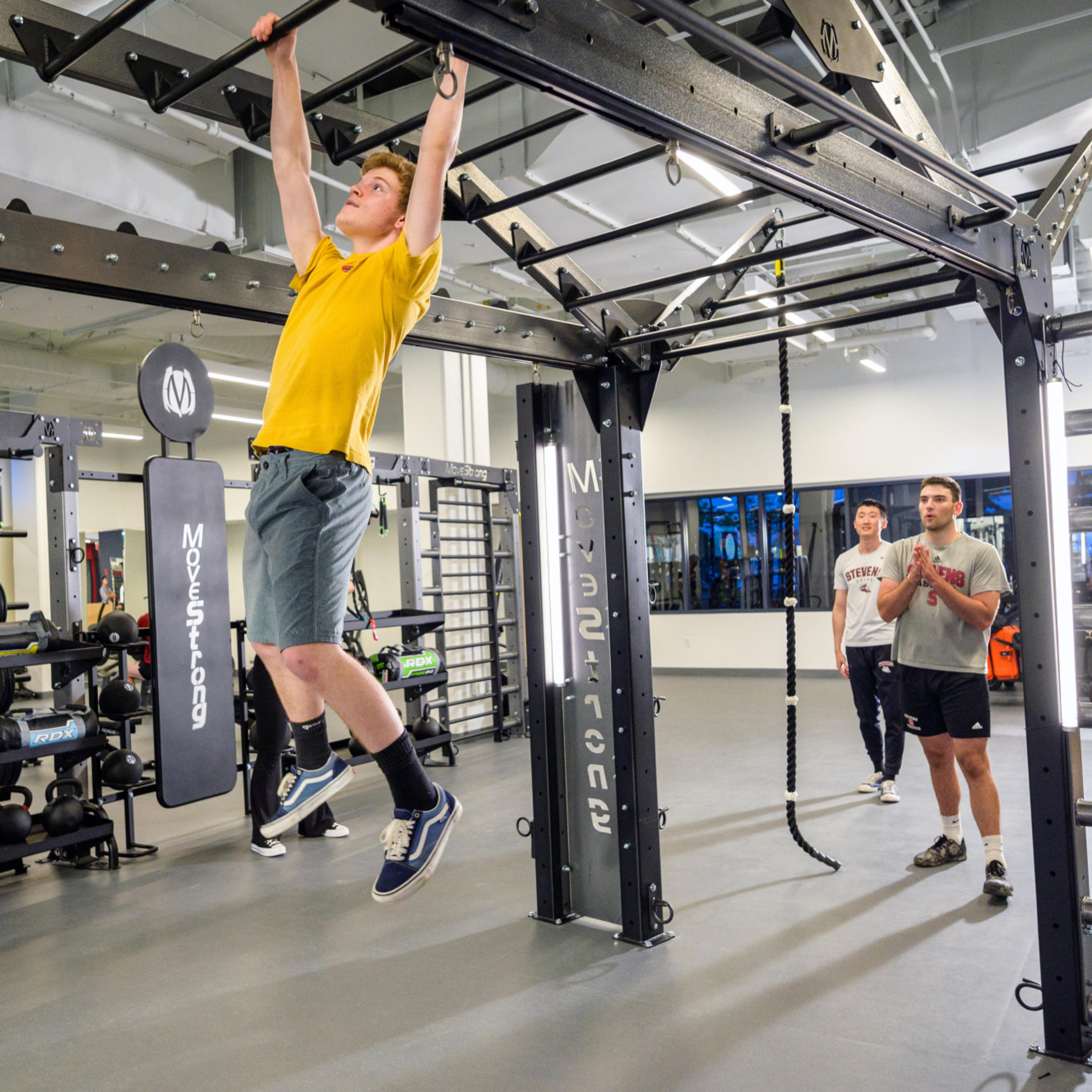 Student in yellow shirt traverses structure while other students cheer him on.