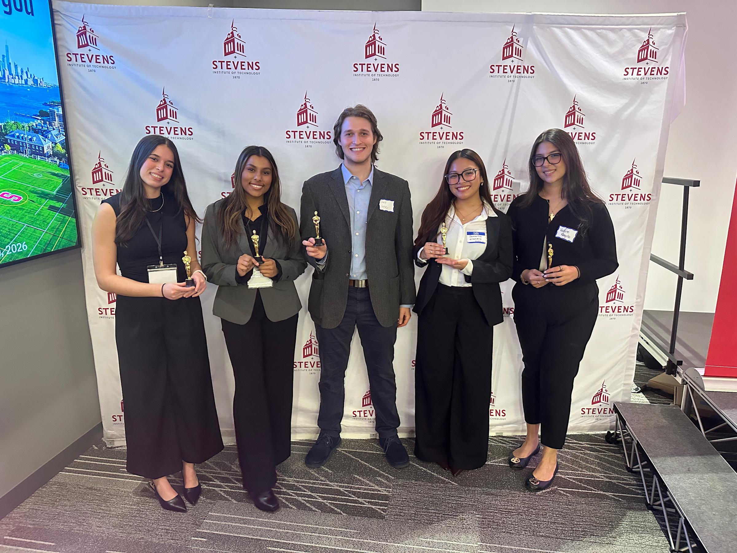 A group of four female students and one male student posing in a row in front of a Stevens Institute of Technology backdrop, each holding an award they received. 
