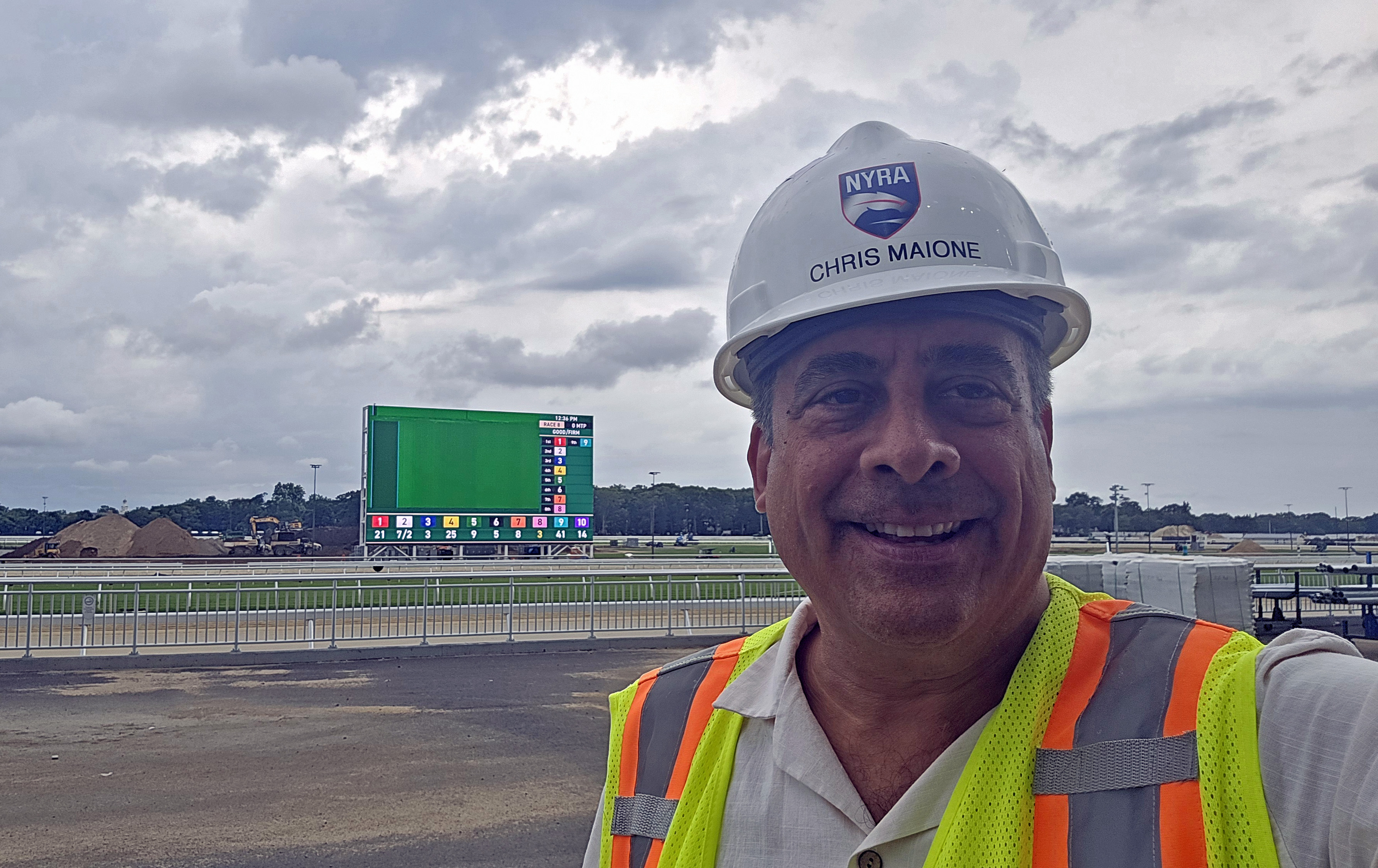 Maione at Belmont Park racetrack in construction hat and vest, with its six-story Infield LED video board that he helped bring to life in the background.