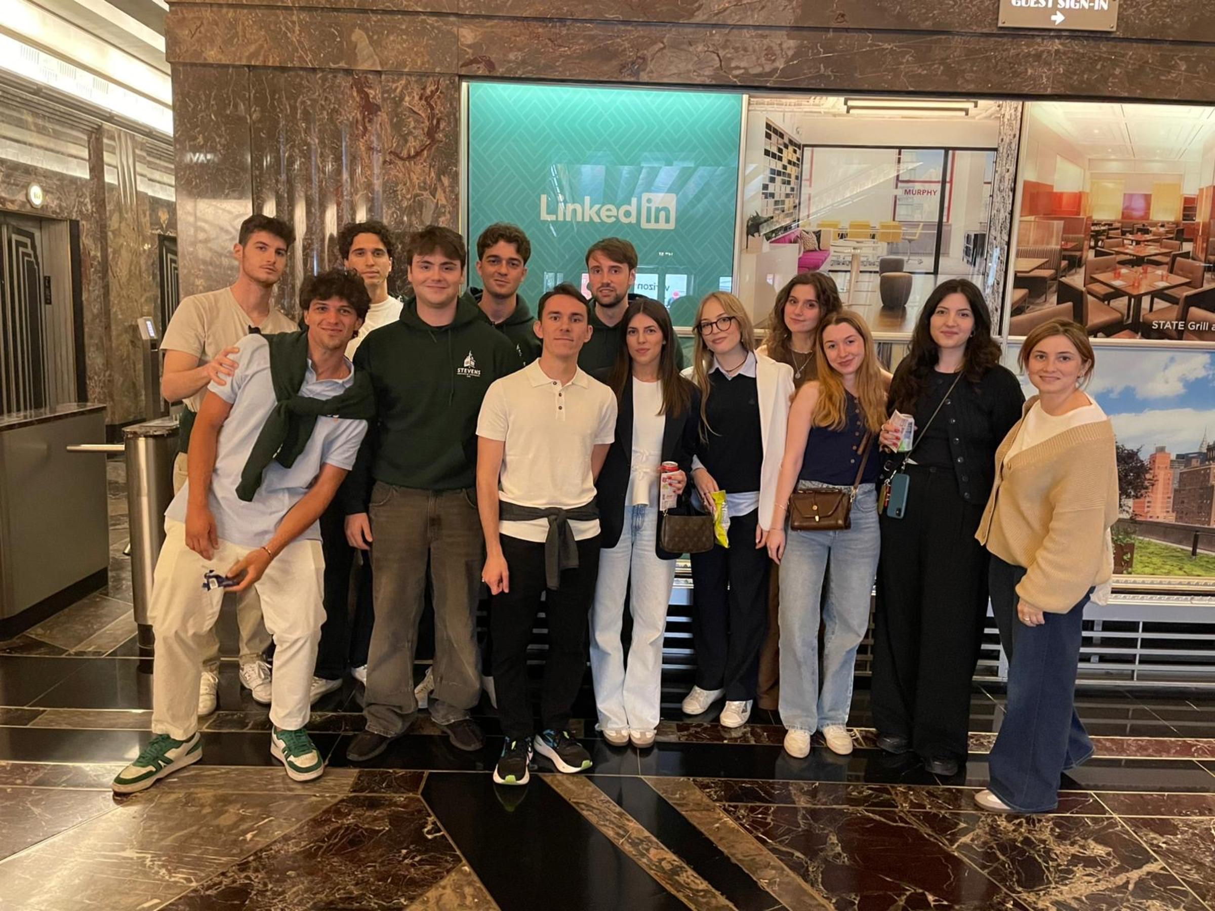 A group of students gathers in a marble-floored office lobby in front of a large LinkedIn logo screen, standing near elevators and glass office spaces during what appears to be a company visit.