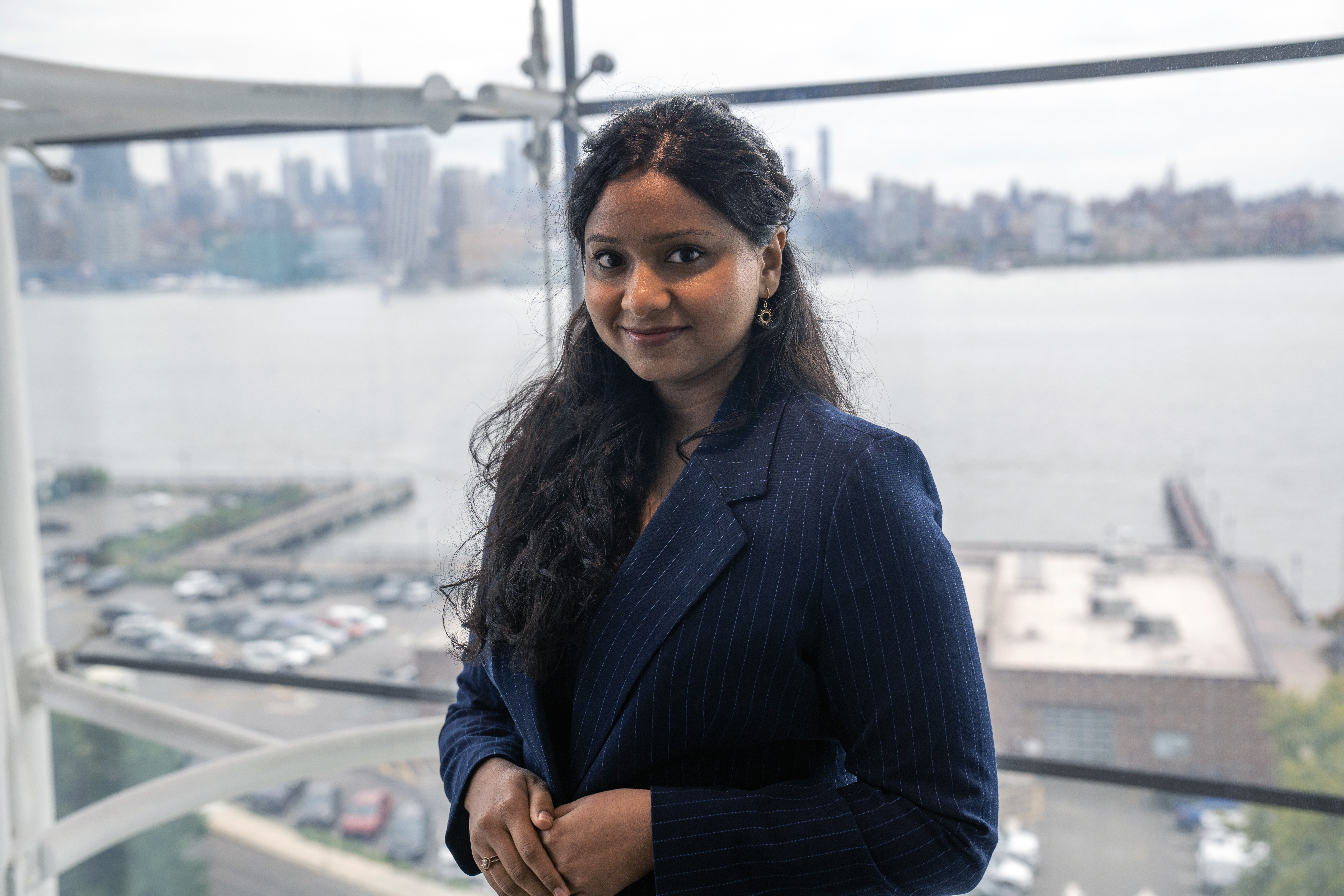 A woman with long dark hair wearing a navy pinstripe blazer smiles at the camera while standing by large windows overlooking a city skyline and waterfront. The professional portrait captures her in what appears to be a high-rise office building.