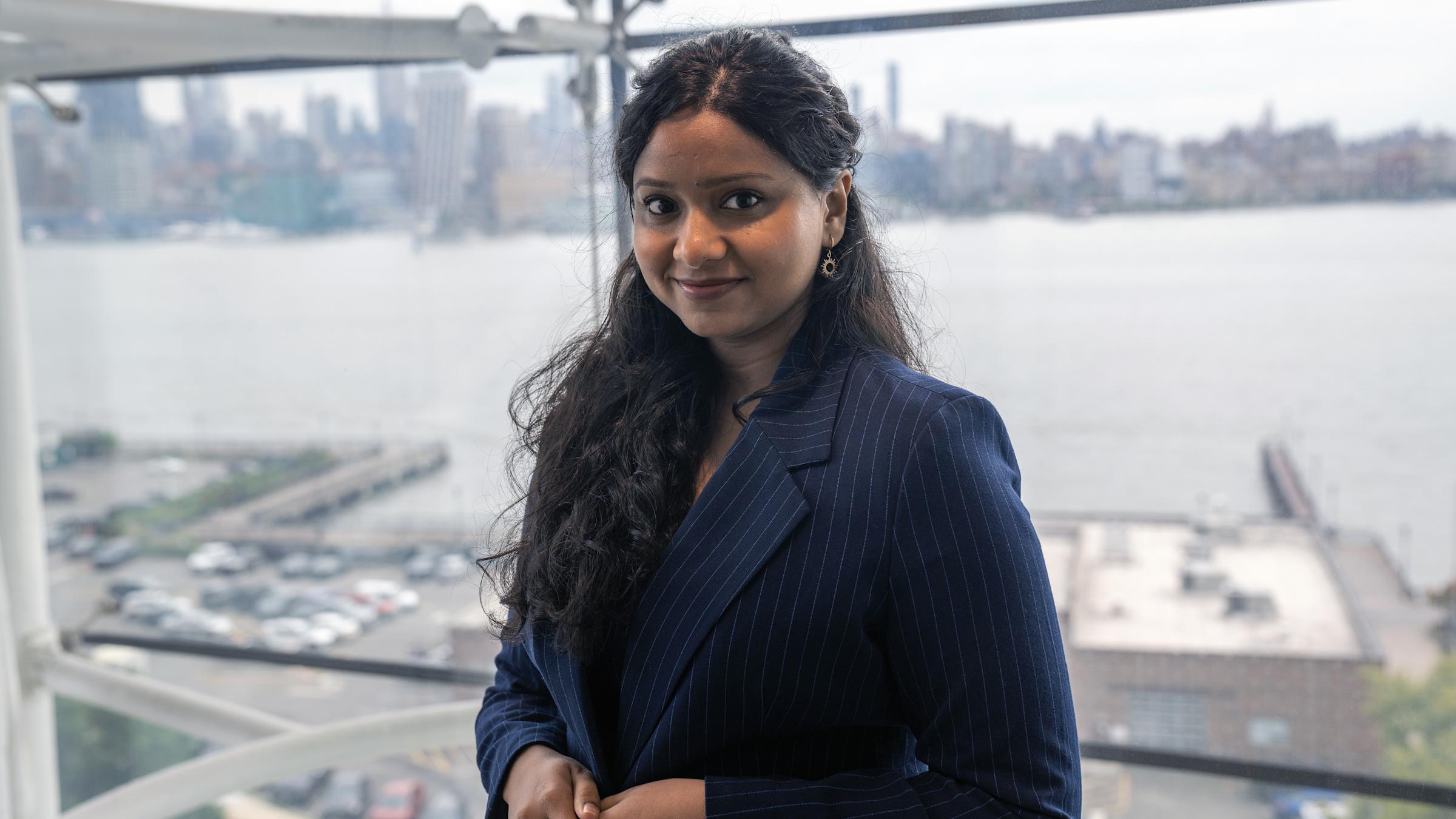 A woman with long dark hair wearing a navy pinstripe blazer smiles at the camera while standing by large windows overlooking a city skyline and waterfront. The professional portrait captures her in what appears to be a high-rise office building.