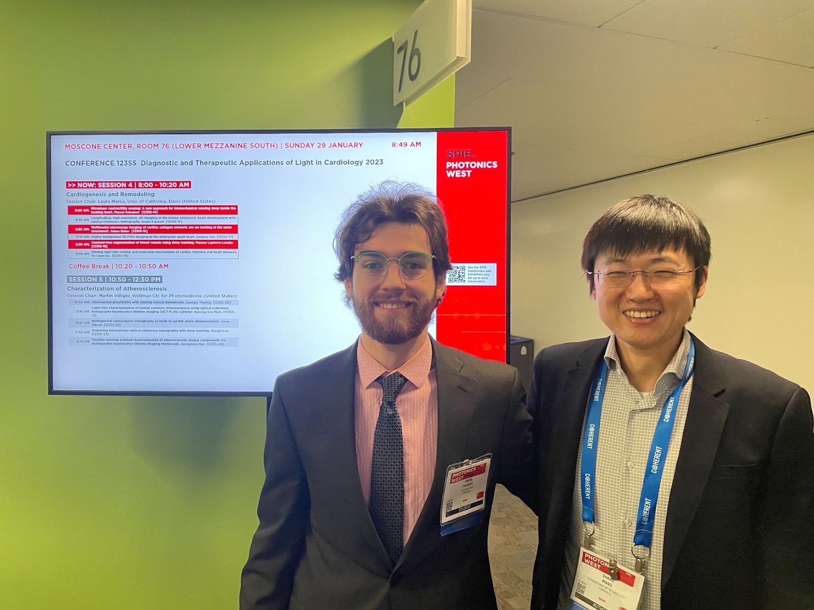 Andre Faubert and Shang Wang standing in front of list of conference proceedings on a television screen