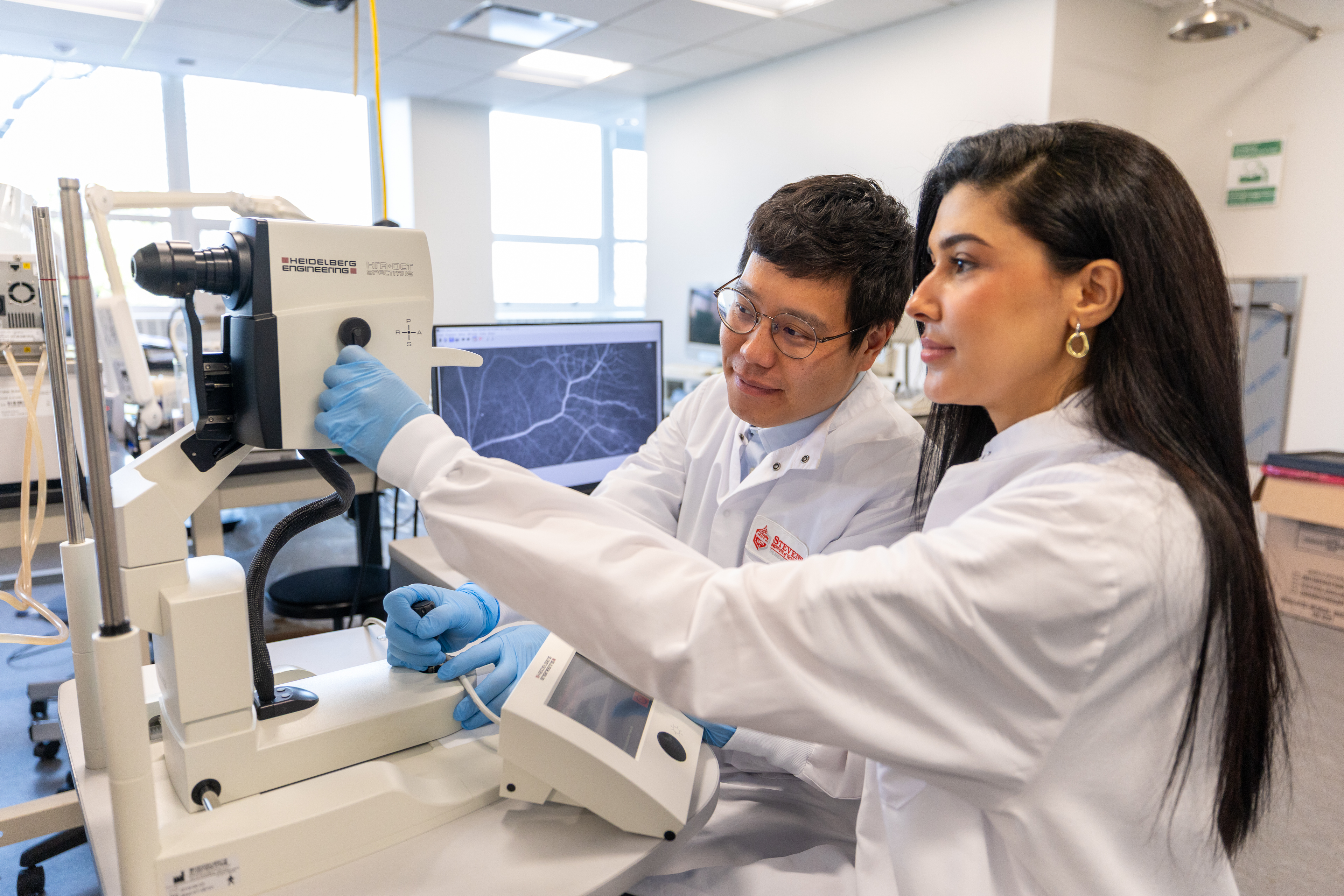 Two graduate students in lab coats work at a piece of lab equipment.