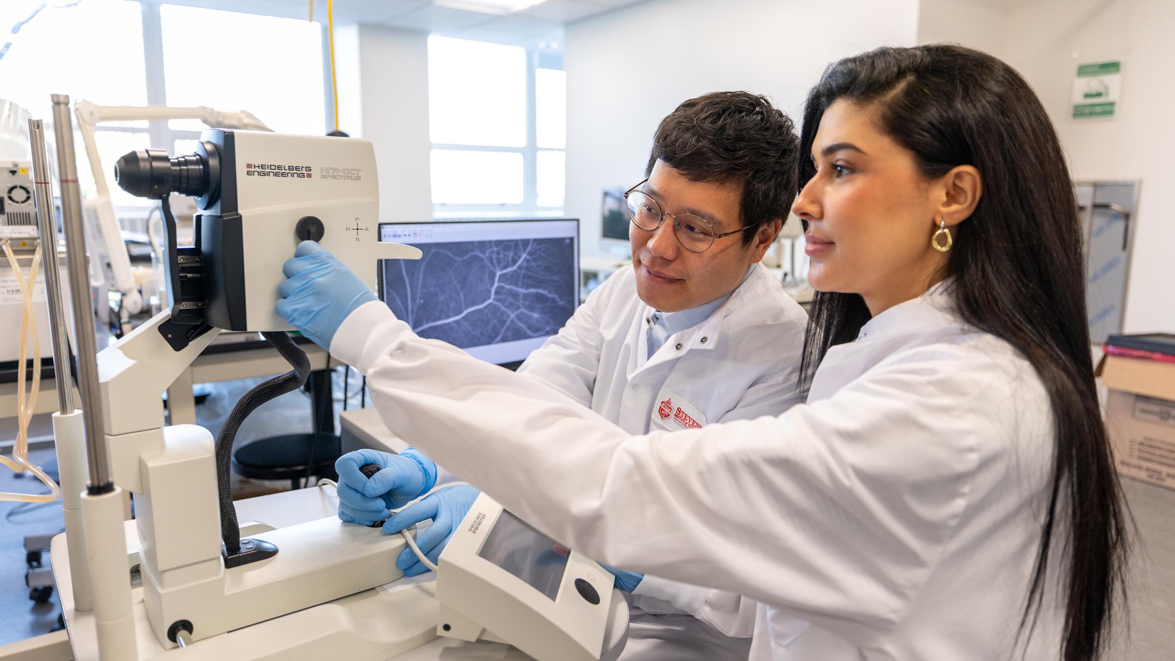Two graduate students in lab coats work at a piece of lab equipment.