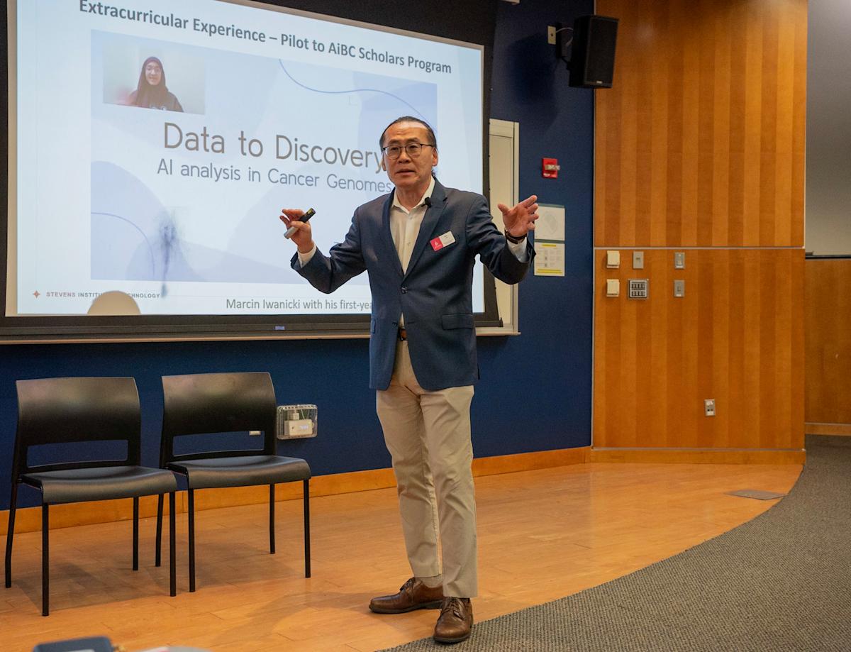 Woo Lee stands in front of a presentation monitor addressing an audience with a microphone.