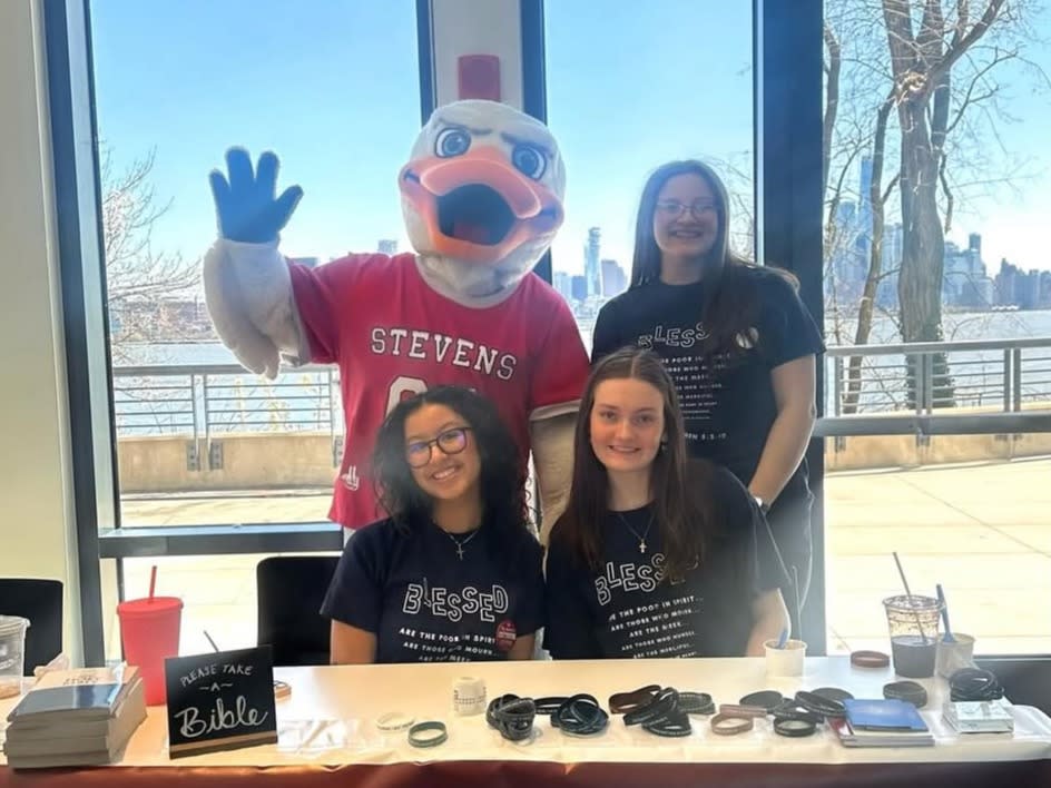 Three smiling students in matching navy "Blessed" T-shirts sit and stand behind a table, joined by a waving duck mascot in a Stevens jersey, with a city skyline visible through the windows.