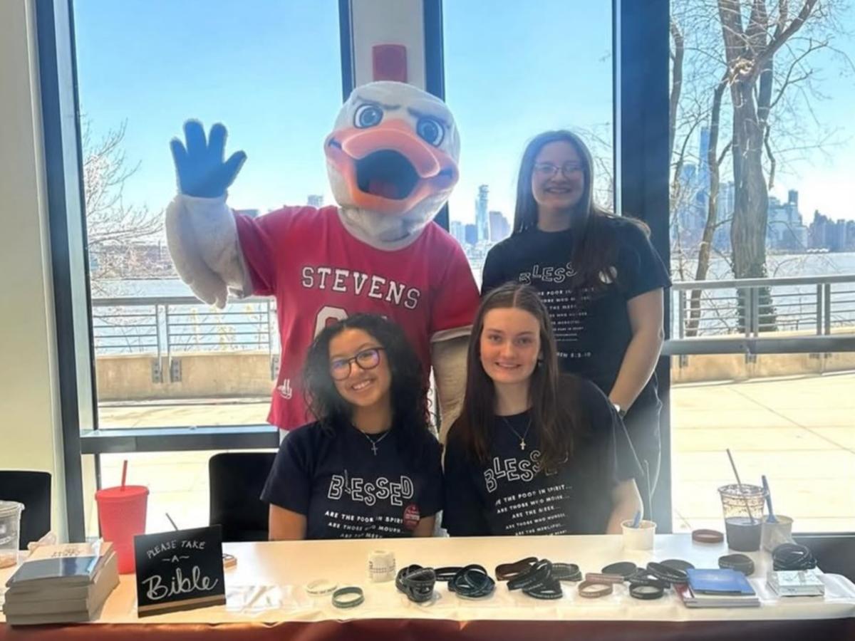 Three smiling students in matching navy "Blessed" T-shirts sit and stand behind a table, joined by a waving duck mascot in a Stevens jersey, with a city skyline visible through the windows.