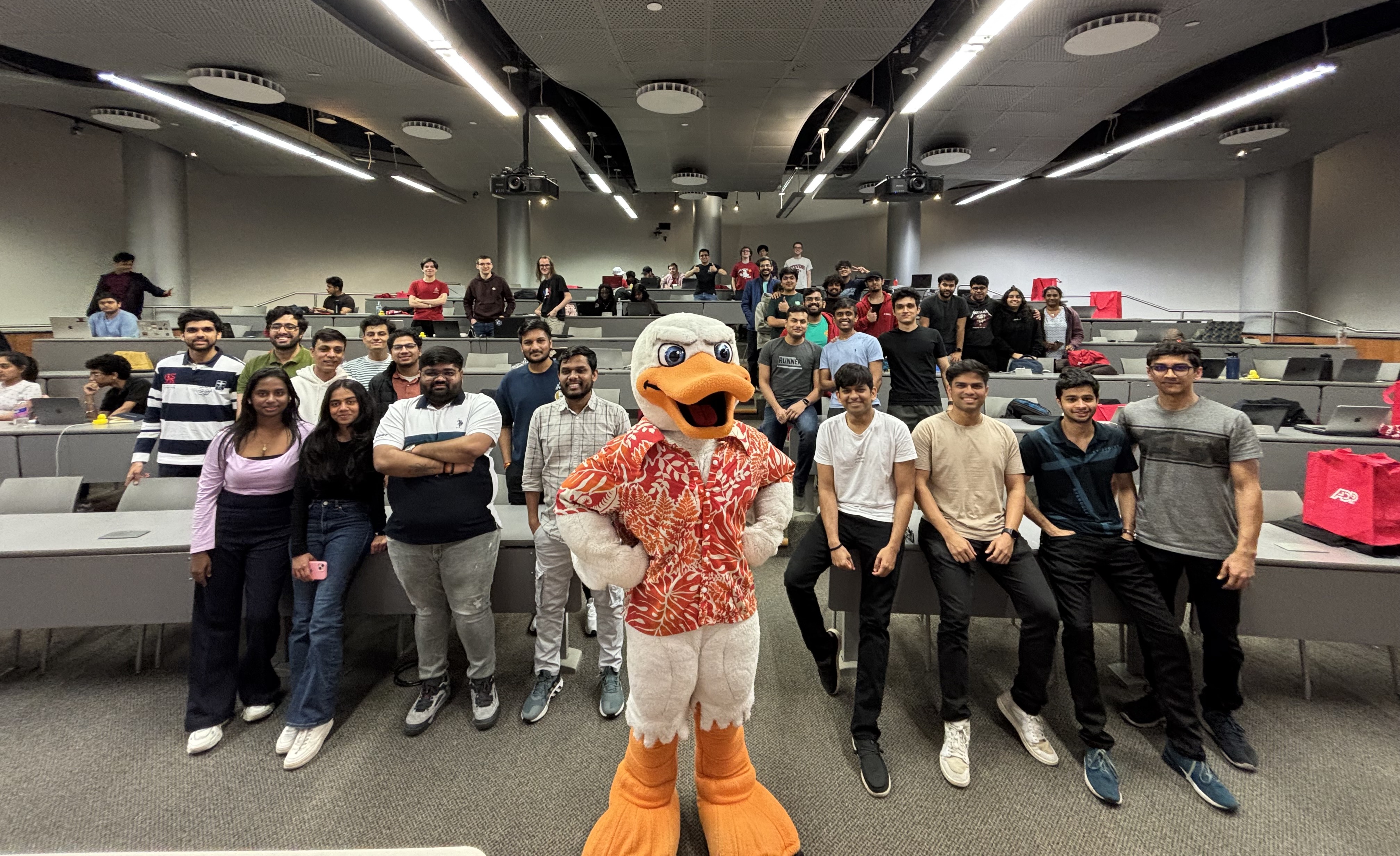 Someone dressed up as the Stevens mascot Atilla the duck stands in the middle of a room, with rows of students  sitting at and on desks behind him.