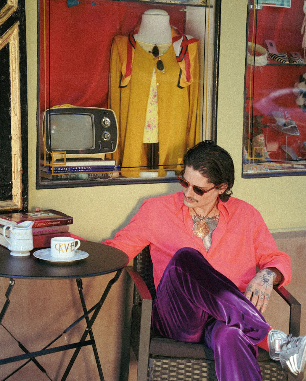 man in sunglasses, shiny purple pants and pink shirt sitting inside a vintage merchandise store, with a display case holding an old TV set, a tunic, scarf, sunglasses and other items.