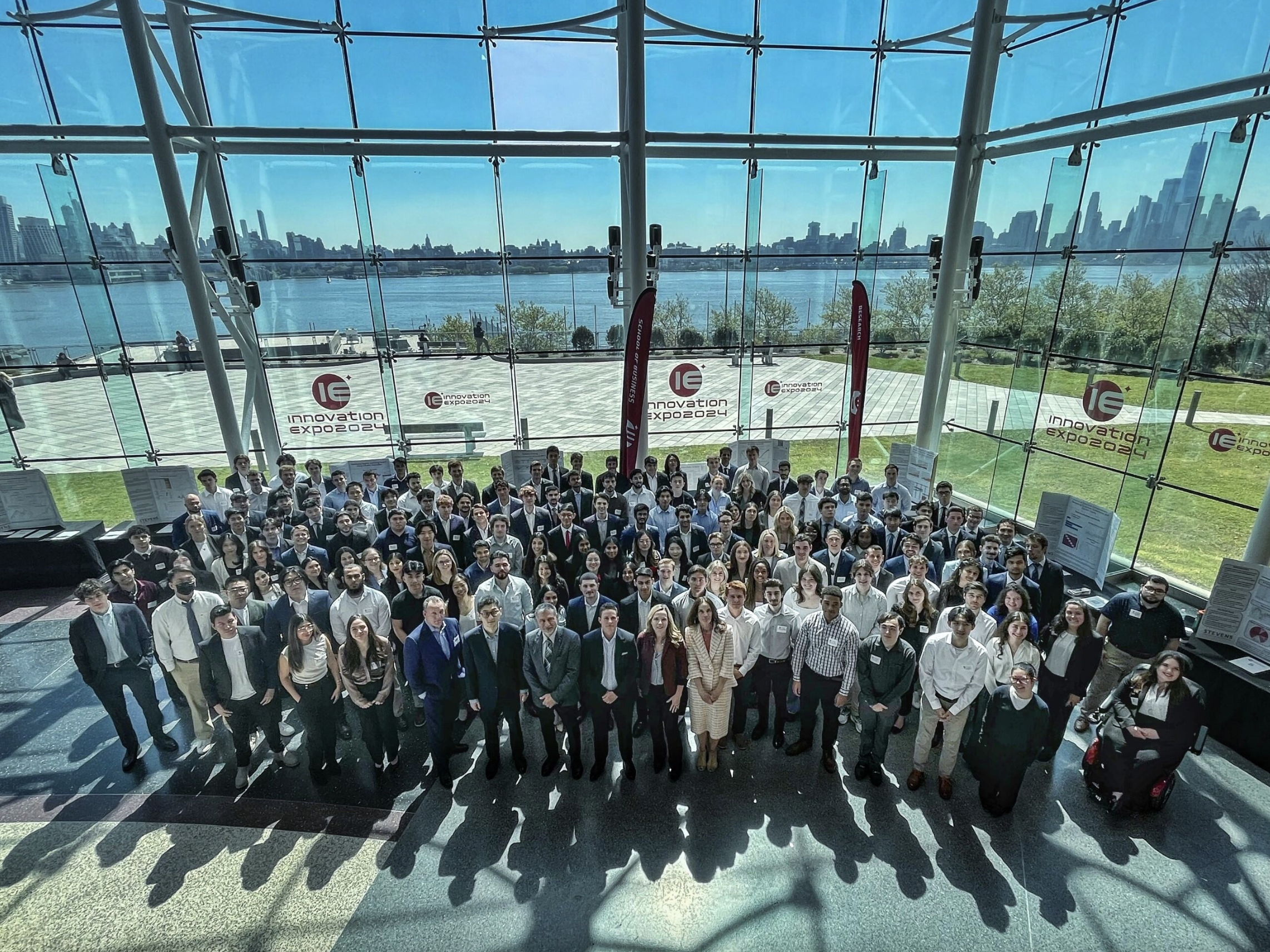 A group shot of the participants in front of the glass atrium walls with the Hudson River and New York City skyline in the background.