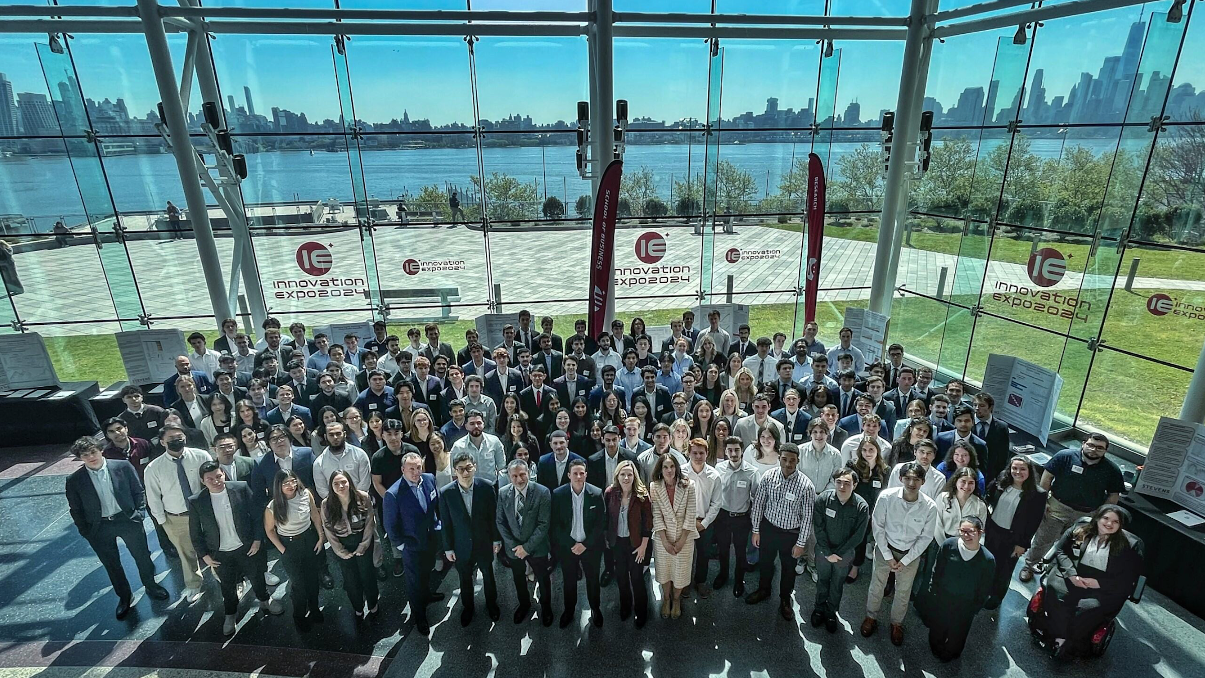 A group shot of the participants in front of the glass atrium walls with the Hudson River and New York City skyline in the background.