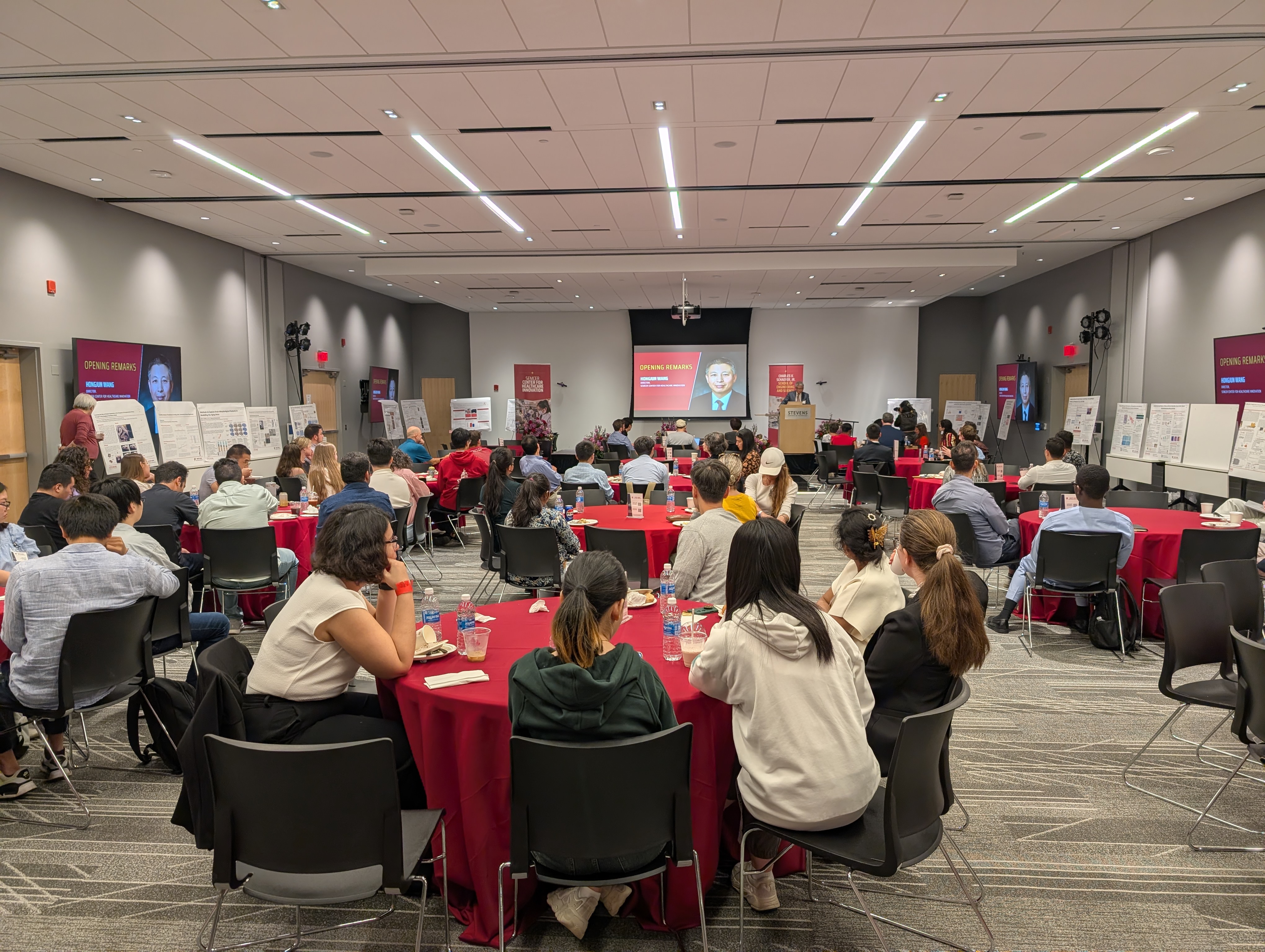 An audience sitting at tables, flanked by poster presentations listening to a speaker at the Next-Gen Healthcare Innovators Symposium.