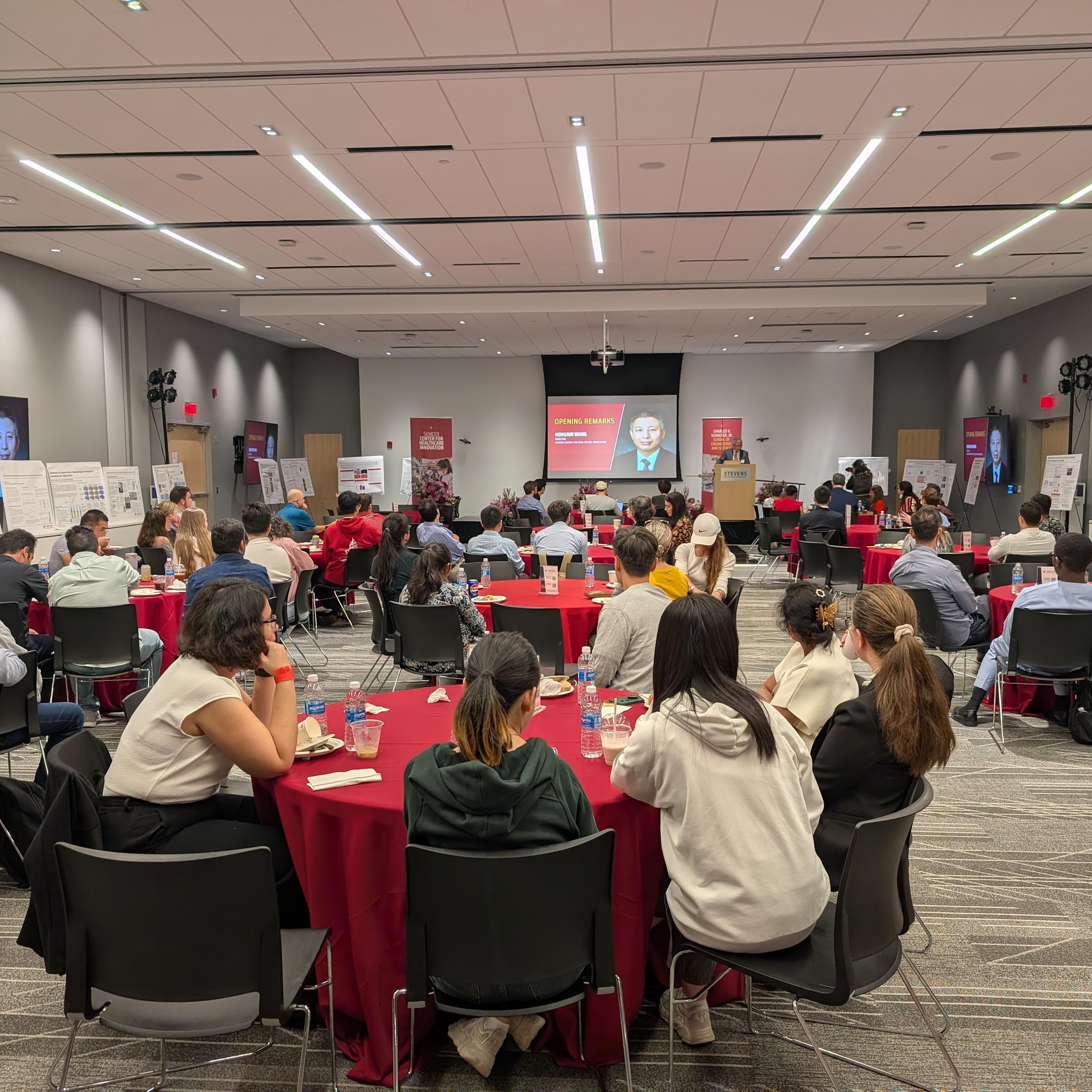 An audience sitting at tables, flanked by poster presentations listening to a speaker at the Next-Gen Healthcare Innovators Symposium.