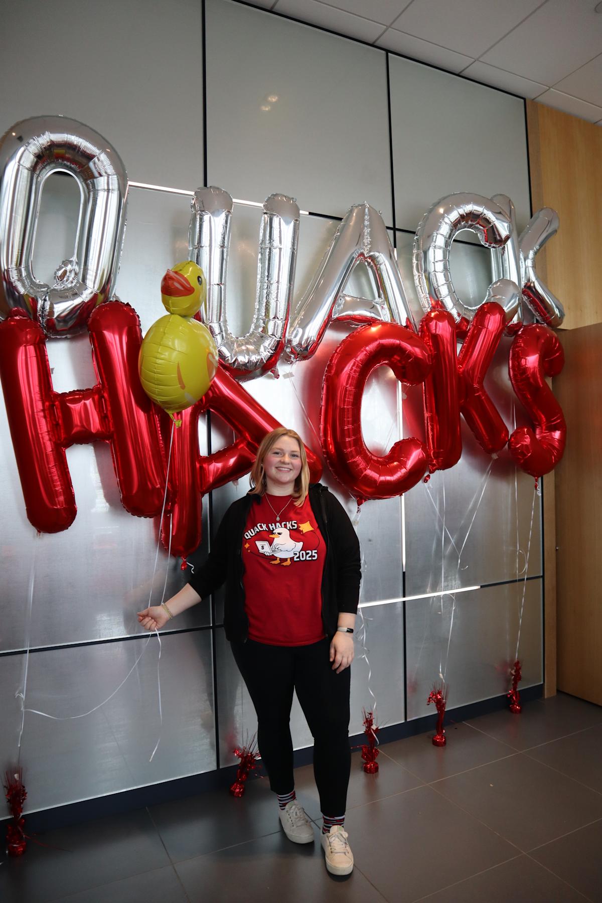 A photo of computer science student Anna Hauk standing in front of balloons that spell out "QUACK HACKS"
