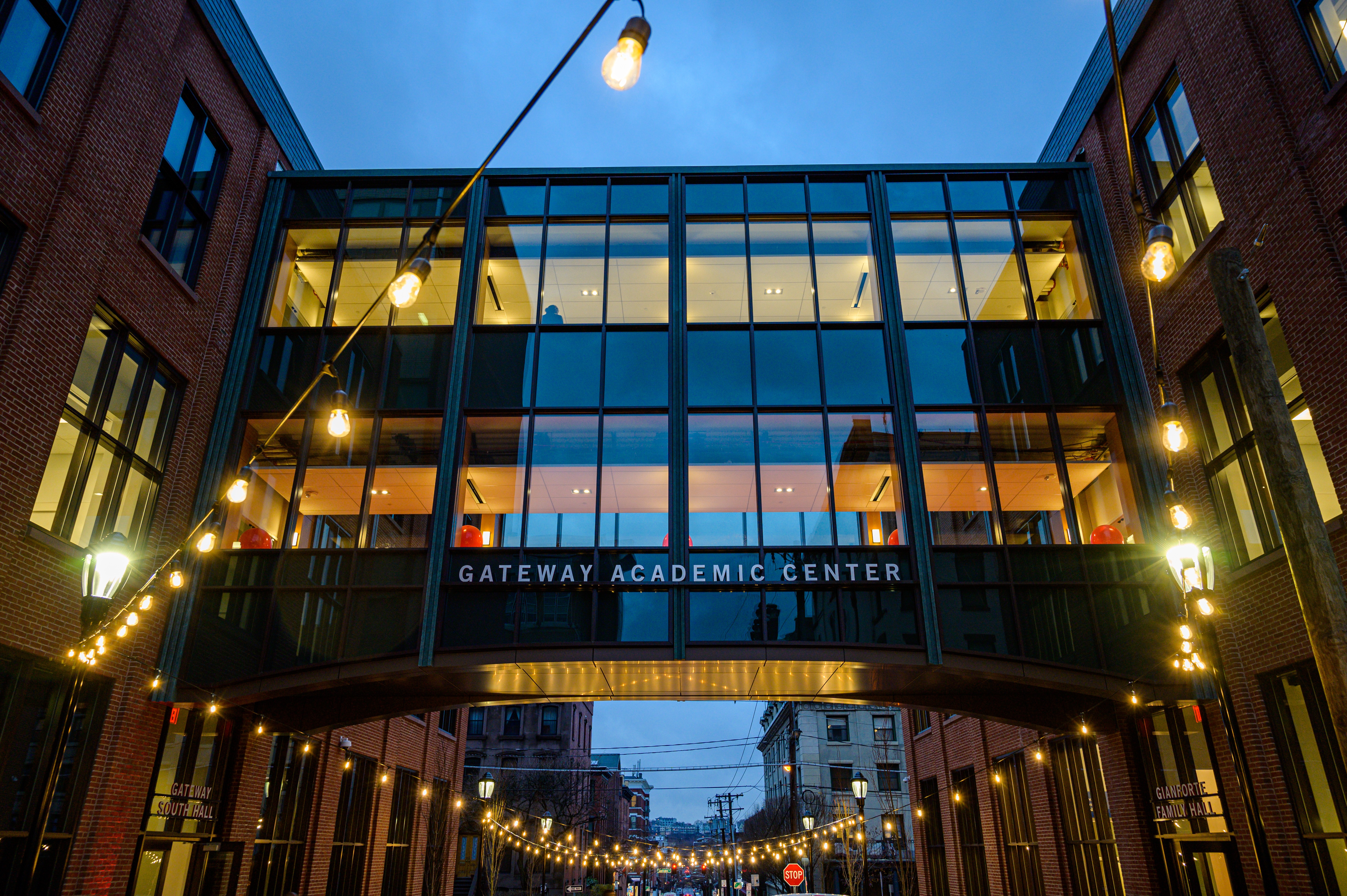 It is nightime. A glass skybridge connecting two building. Lights are strung above.