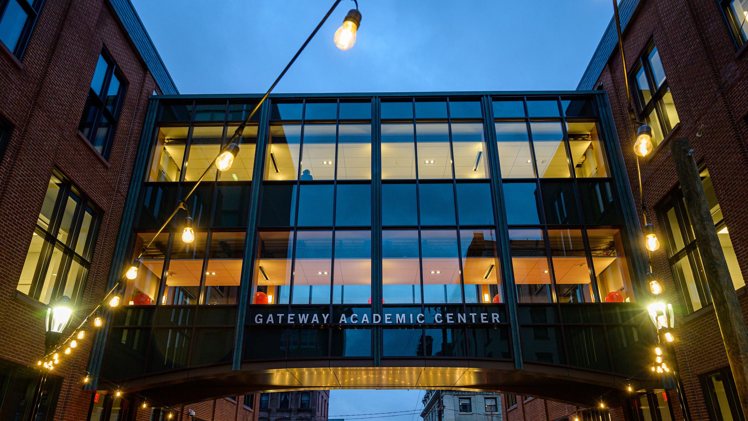 It is nightime. A glass skybridge connecting two building. Lights are strung above.