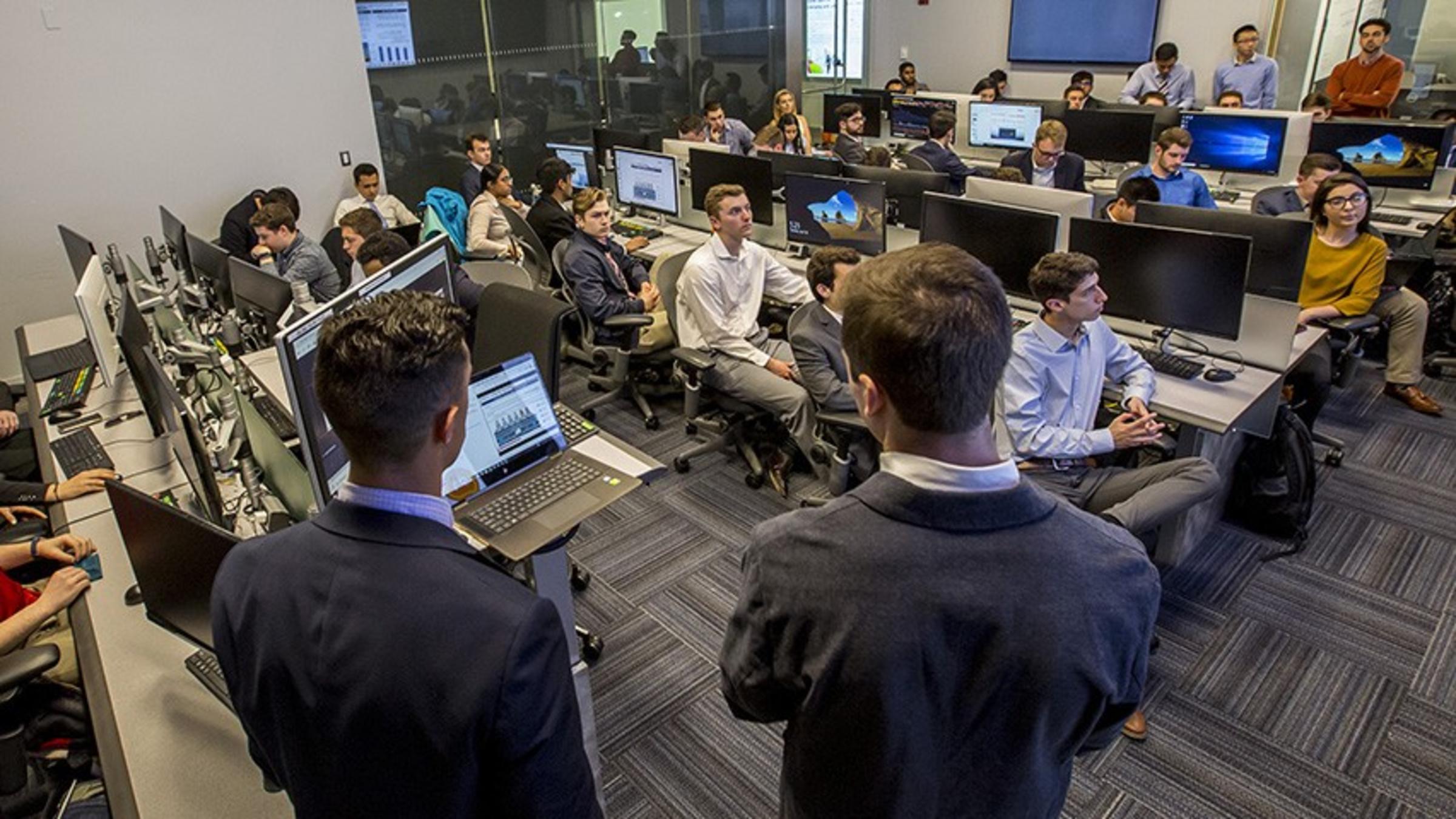 A class in a Bloomberg finance lab being led by two male students wearing dark suits.